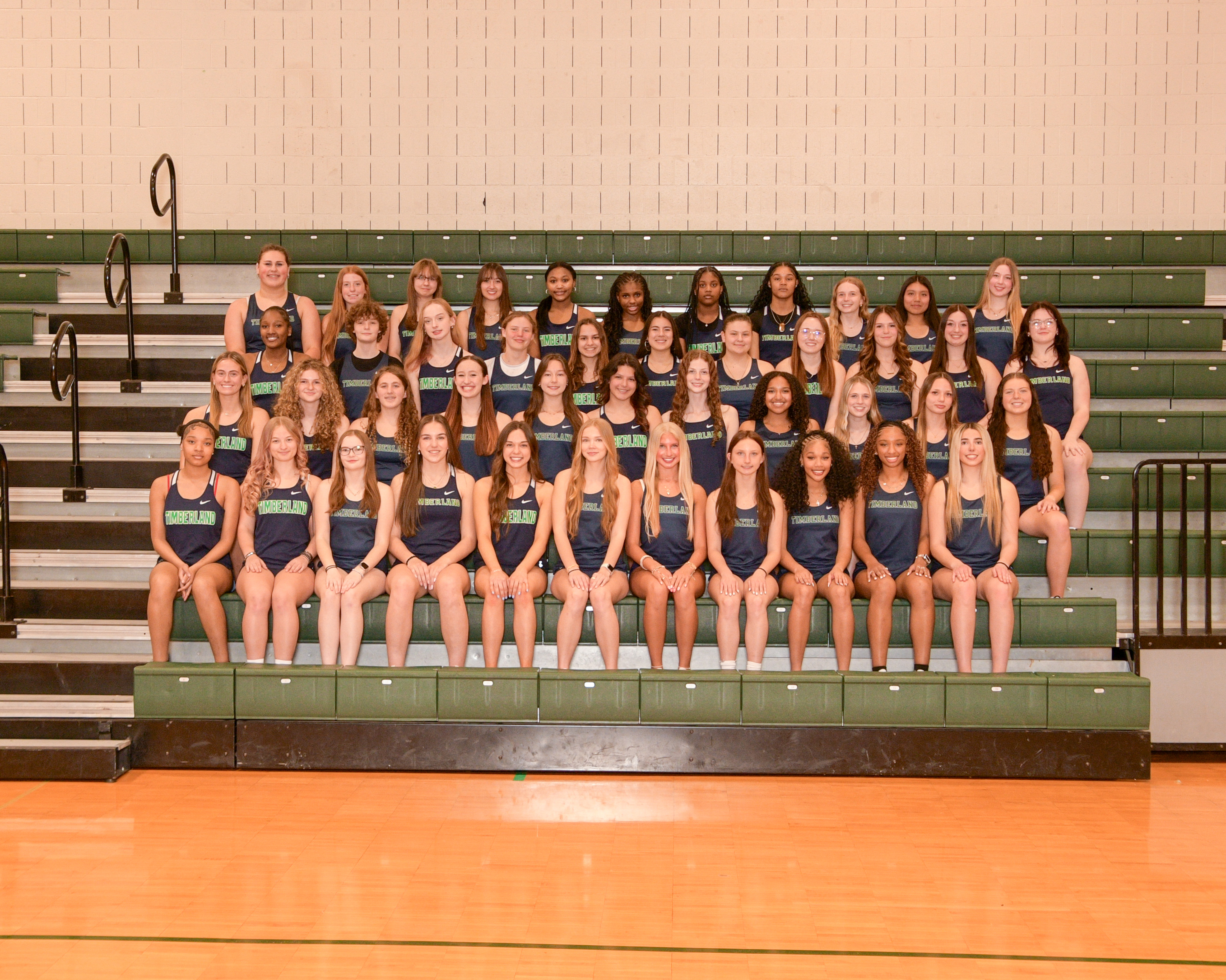 A girls track and field team, posing together on a basketball court with bleachers in the background.