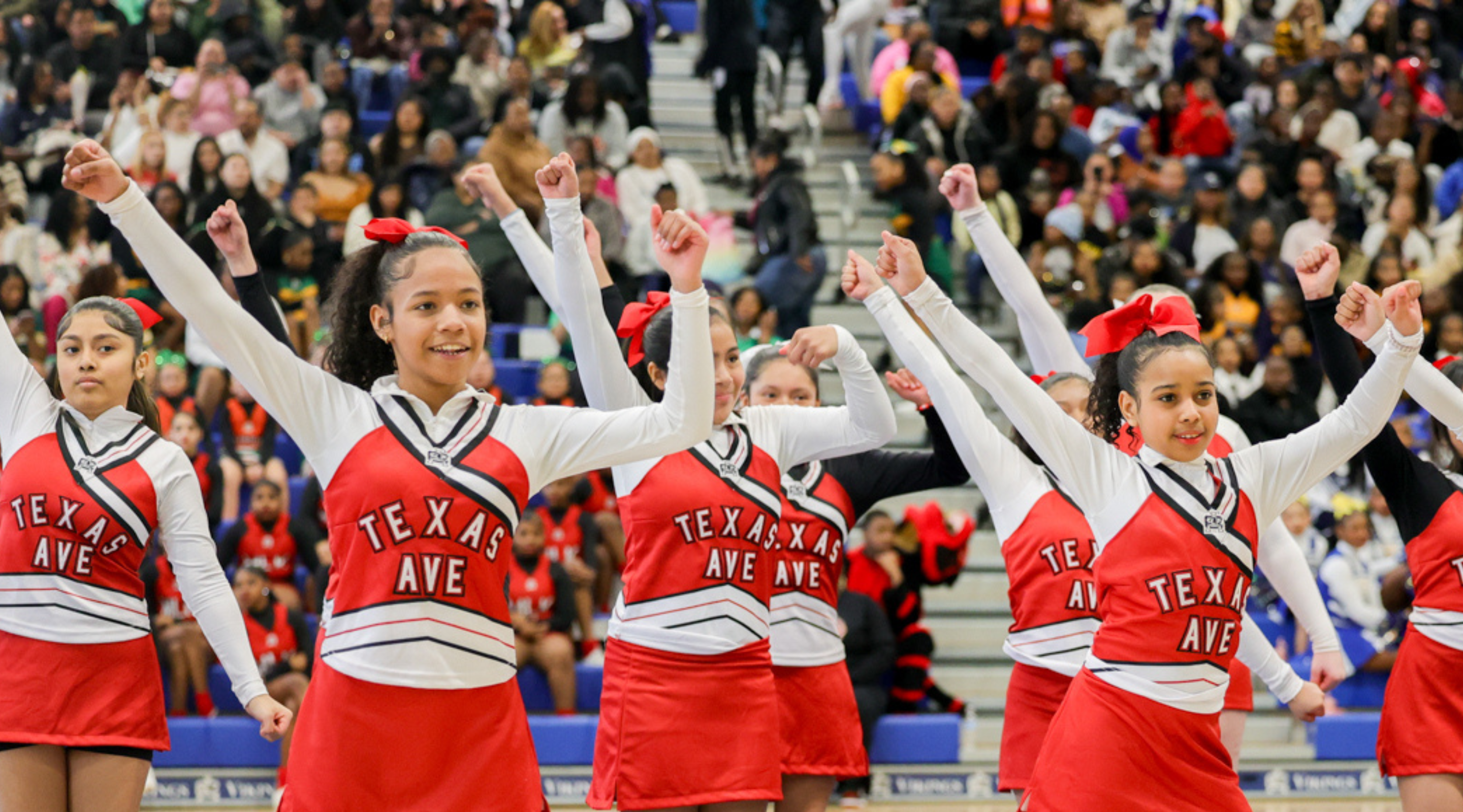 A group of cheerleaders in red uniforms with the text "Texas A&amp;E" performing synchronized dance moves in front of a large crowd.