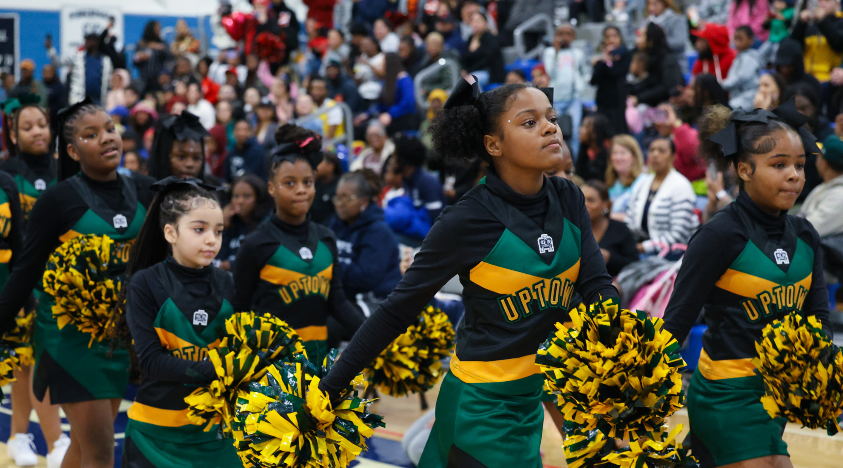 A large crowd of cheerleaders in green and yellow uniforms performing on a stage, with a backdrop of more people in the background.