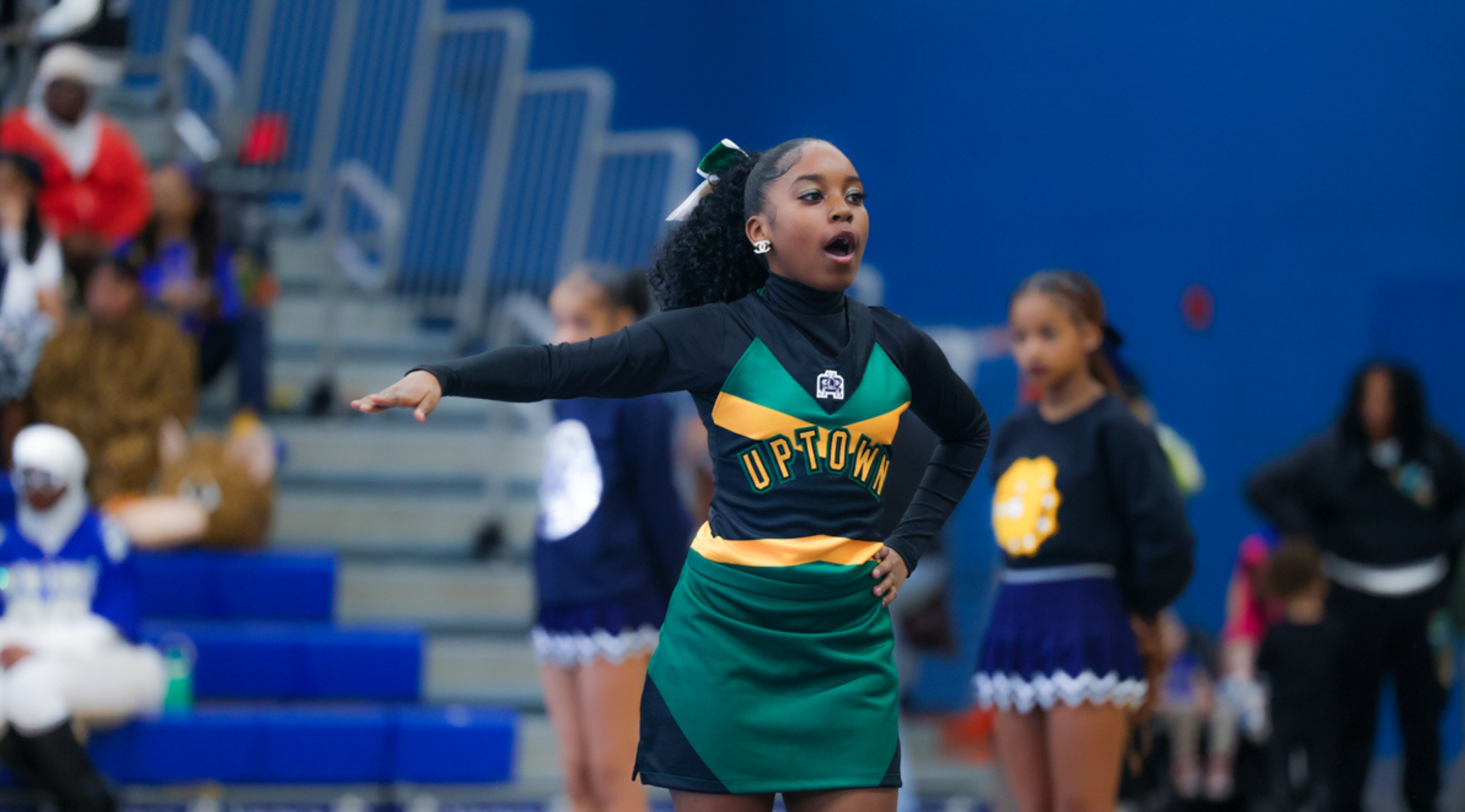 A young woman in a green and yellow cheerleading uniform is performing on a stage with a blue background and spectators in the stands.