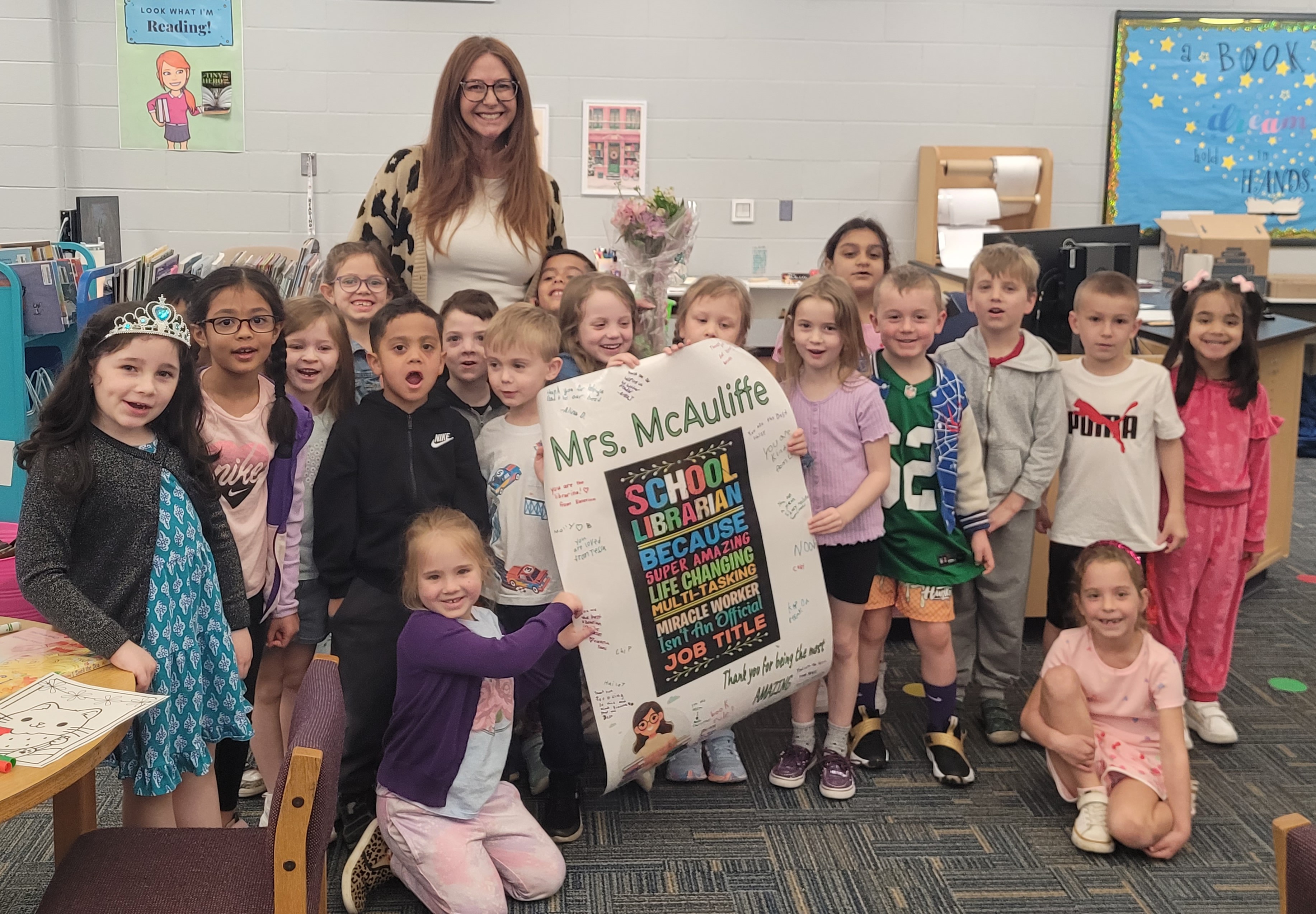The image shows a group of young students, both boys and girls, posing with their teacher in a classroom setting. The teacher is holding a sign that appears to be related to the classroom or school activities.