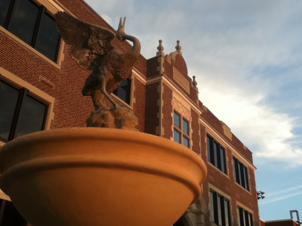 A bronze statue of a goose with its wings spread, perched atop a large stone pedestal in front of the historic Morristown East High School brick building.
