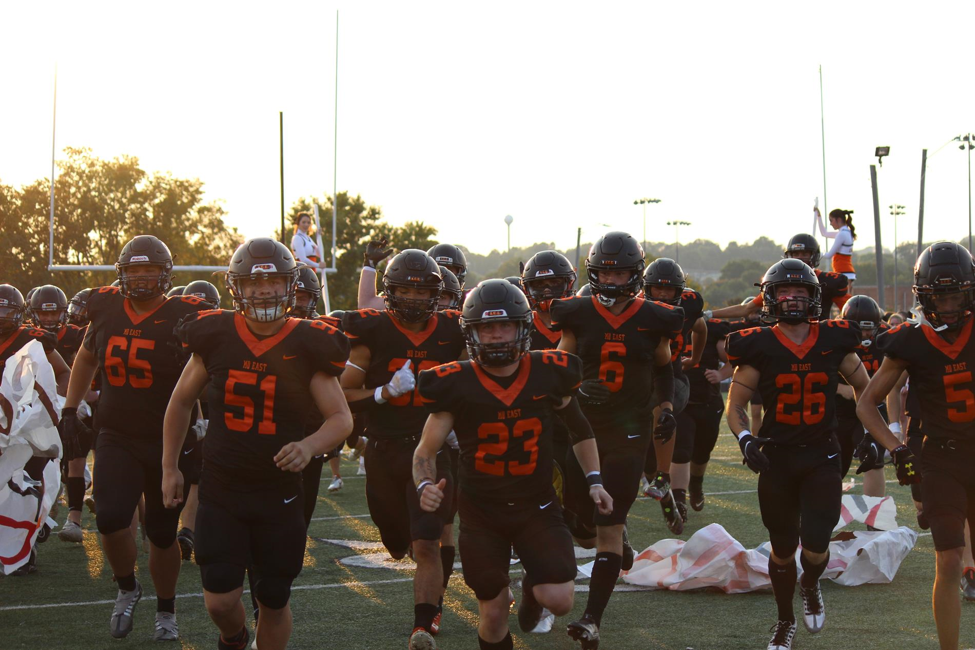 Morristown East Hurricanes football team in black and orange jerseys, running onto the field before a game at sunset. Visible jersey numbers include 65, 51, and 23.