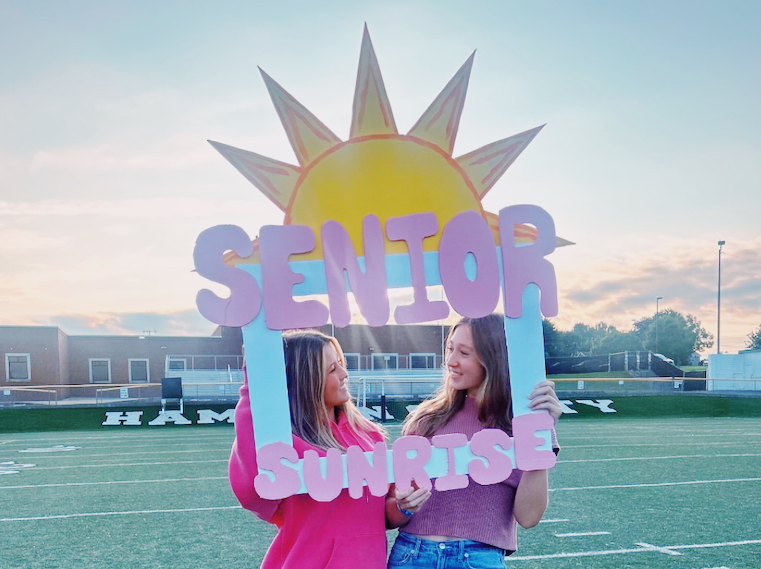 Two female students smiling and holding a decorative photo frame that reads 'Senior Sunrise' on a football field. A large hand-painted sun is in the background as the sun rises over the campus.