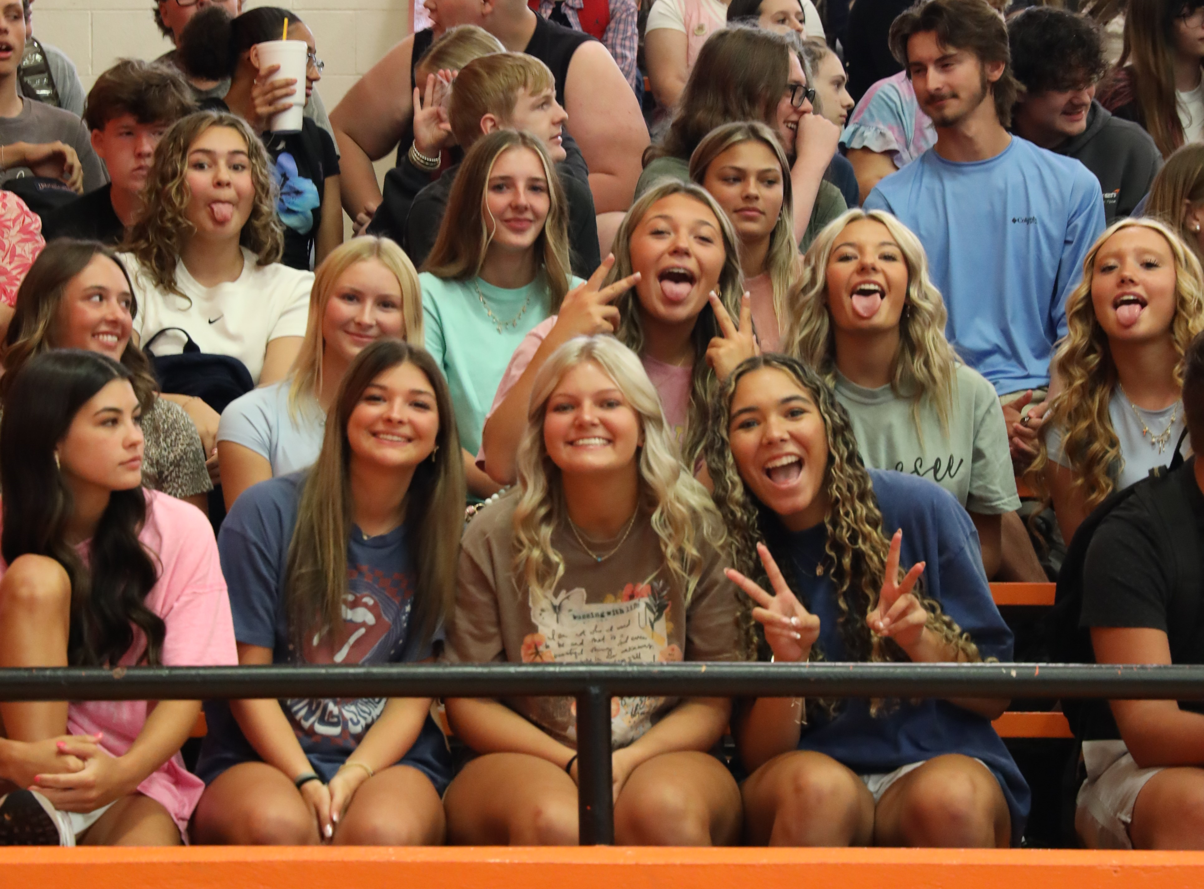 A large group of high school students cheering in the bleachers during a school assembly or pep rally. Several students in the front row are smiling, making peace signs, and showing school spirit.