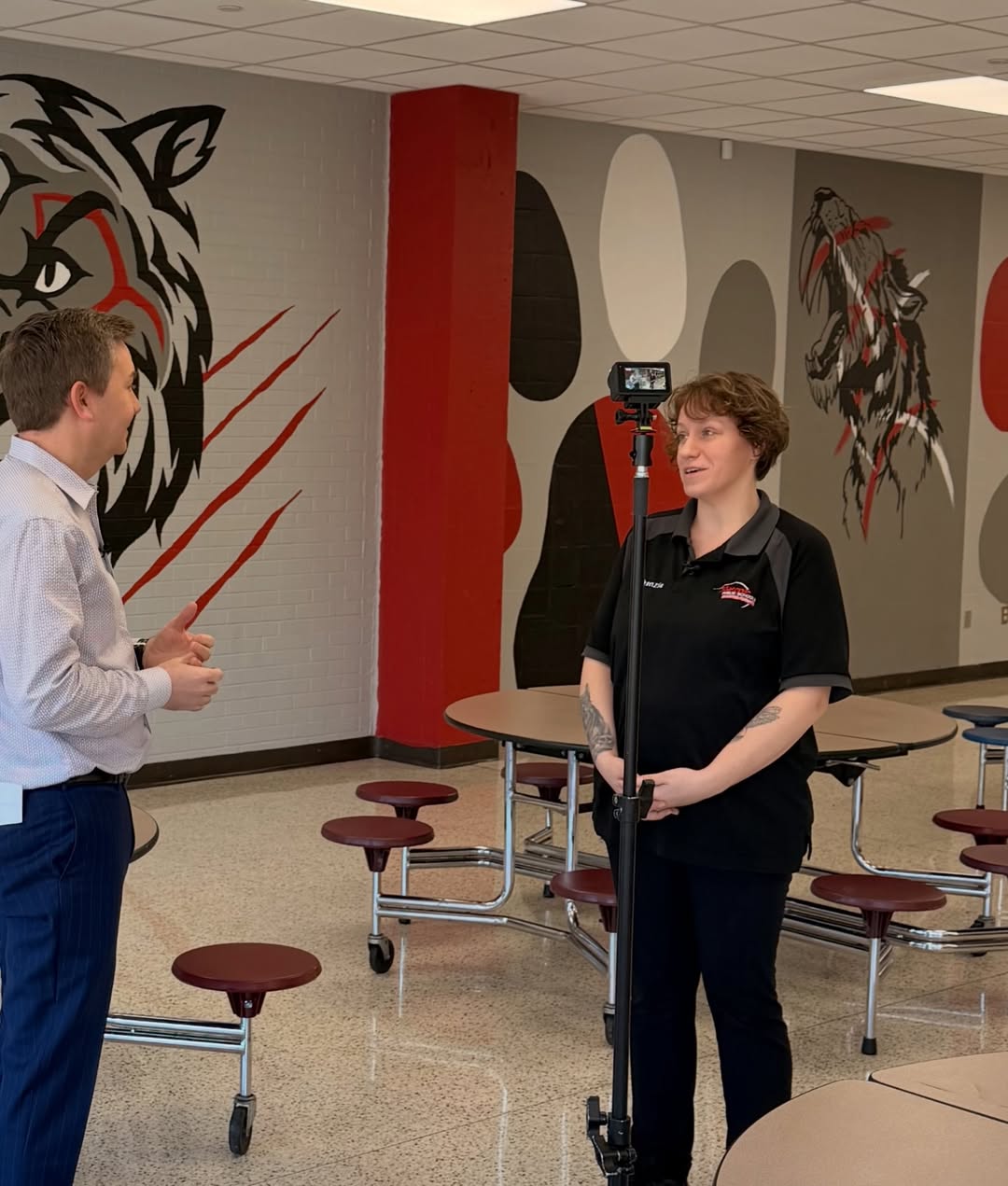 A man in a light blue shirt and dark pants speaks to a woman in a black polo shirt and dark pants, with a camera on a tripod between them, in a cafeteria with tiger murals on the walls.