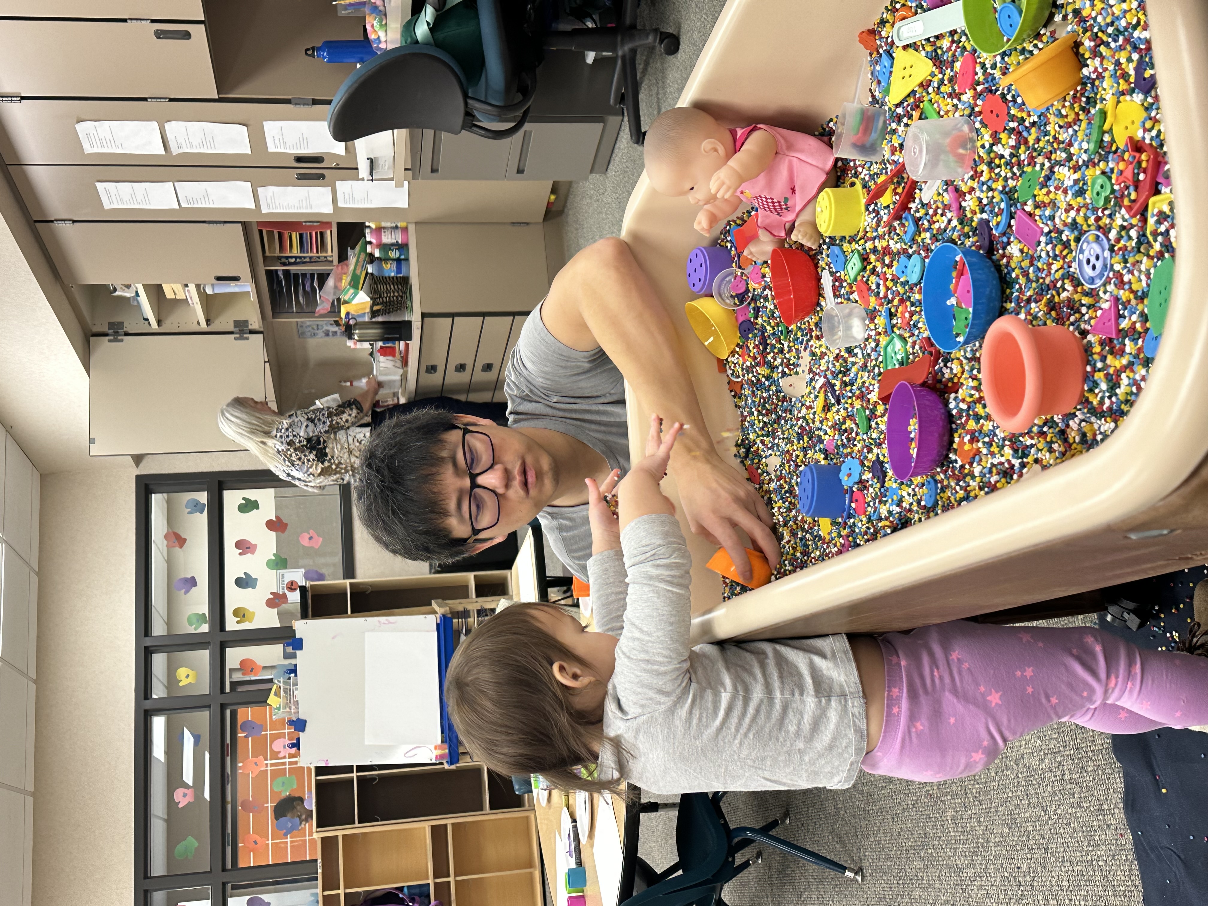 A classroom setting with a teacher and a young student engaged in play with a variety of colorful toys and objects scattered on the floor.