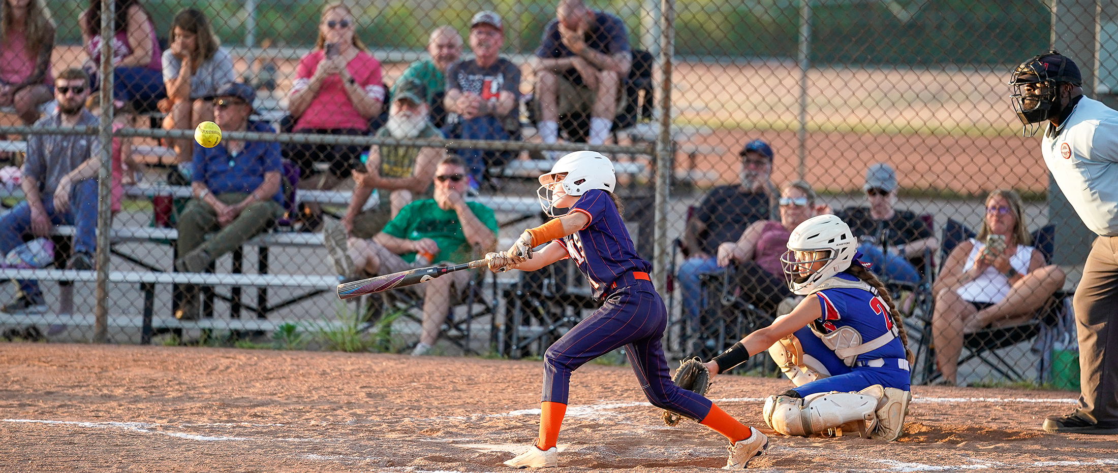 The image shows a softball game in progress, with a player in a purple uniform batting and a catcher in a blue uniform crouched behind home plate, while a crowd of spectators watches from the bleachers in the background.