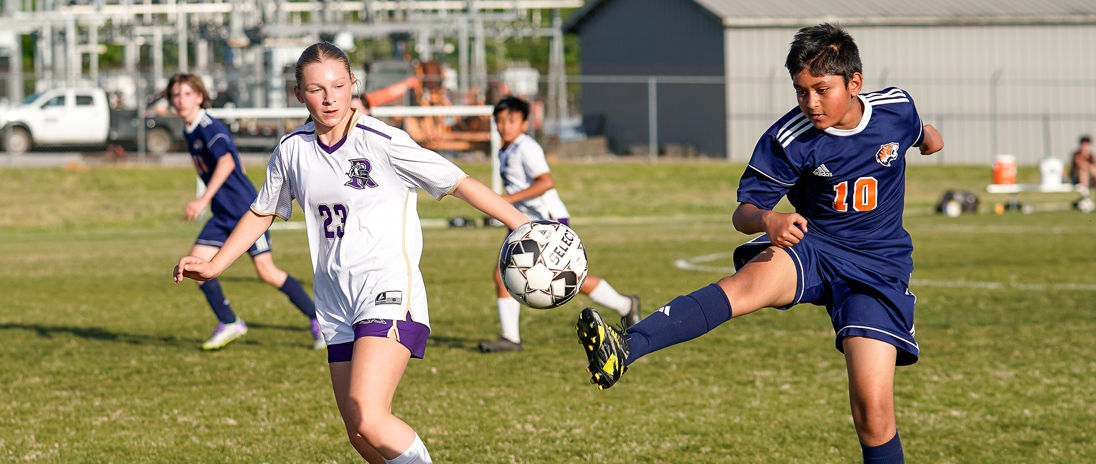 Two soccer players, one in a white jersey and the other in a blue jersey, are competing for the ball on a grassy field, with other players visible in the background.