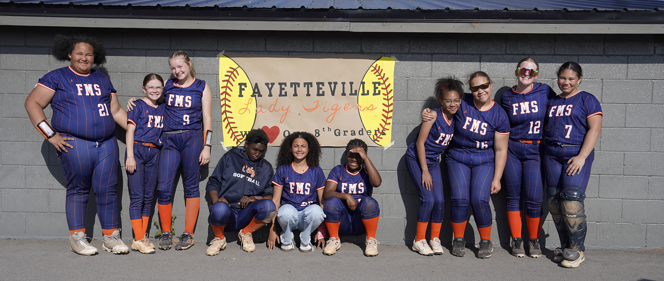A group of young female softball players in uniform standing in front of a wall with the text "Fayetteville" and a softball graphic.