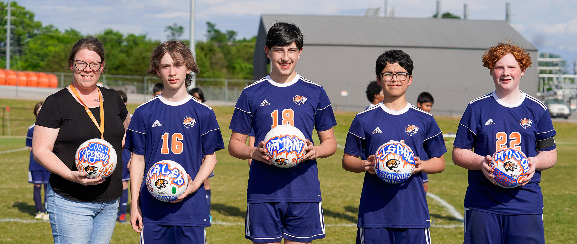 A group of young soccer players in blue uniforms stand on a field, with adults in casual clothing standing behind them.