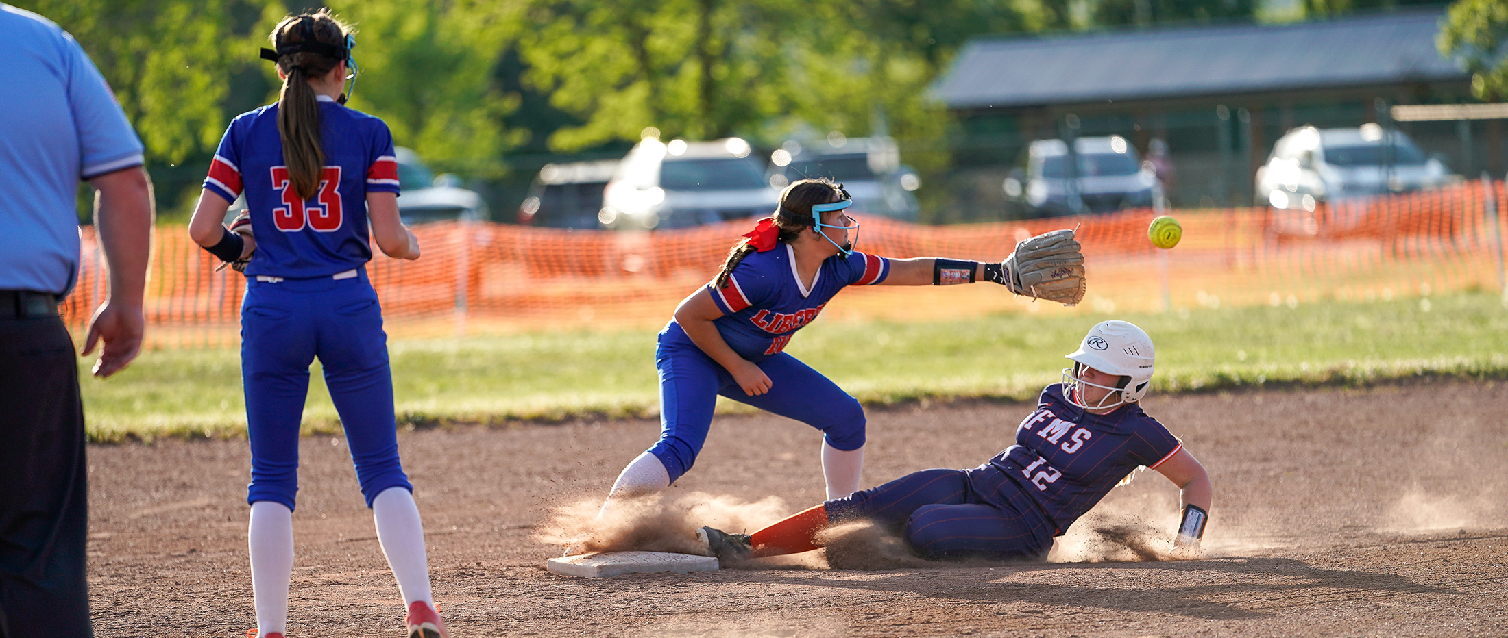 The image depicts a softball game, with a player in a blue uniform sliding into a base as another player in a blue uniform prepares to catch the ball, while a third player in a blue uniform stands in the background.