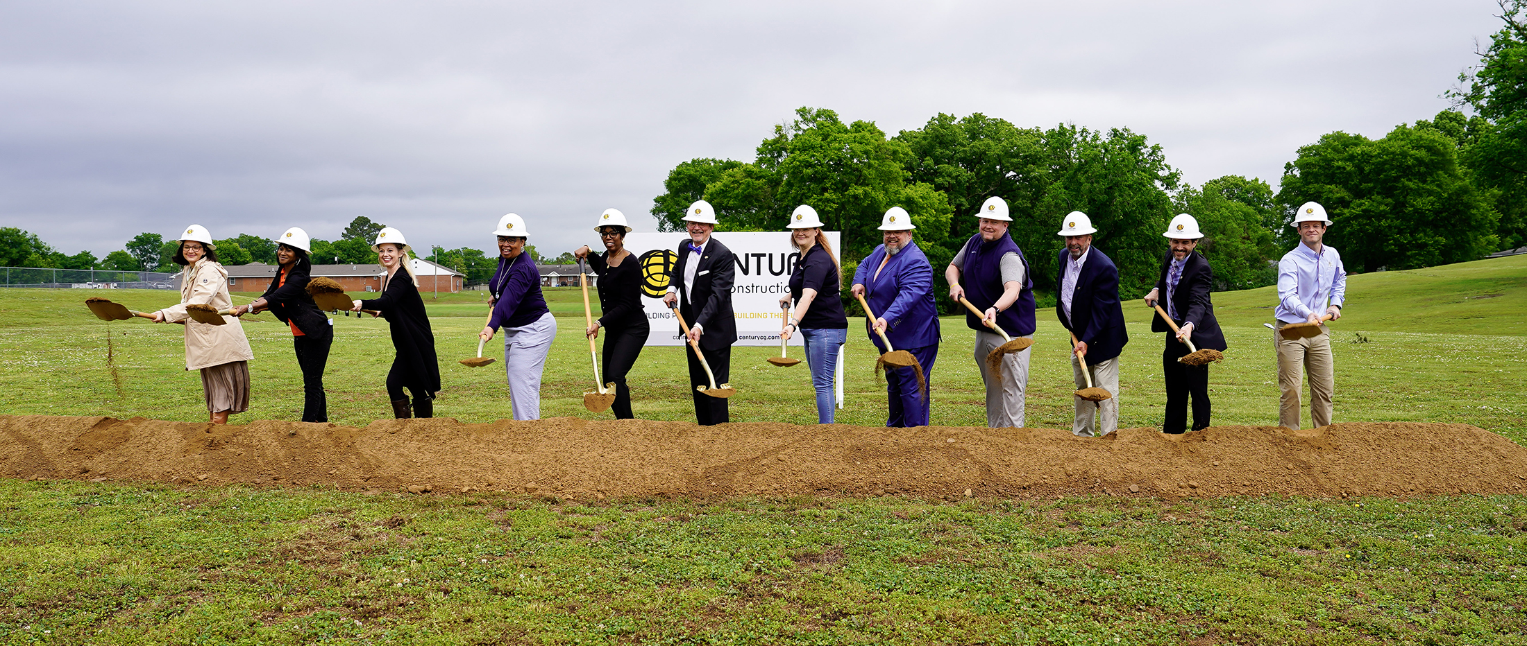 A group of people in various outfits, including hard hats, standing in a field with trees and a cloudy sky in the background.