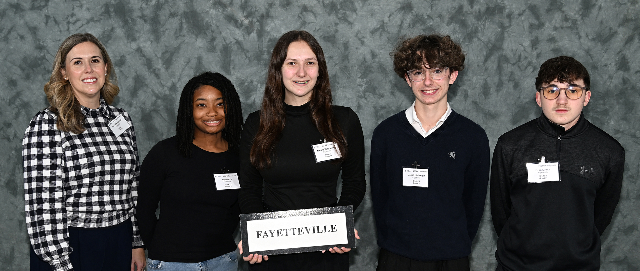 The image shows a group of five people, all wearing black or checkered shirts, standing in front of a gray background and holding a sign that says "Fayetteville".