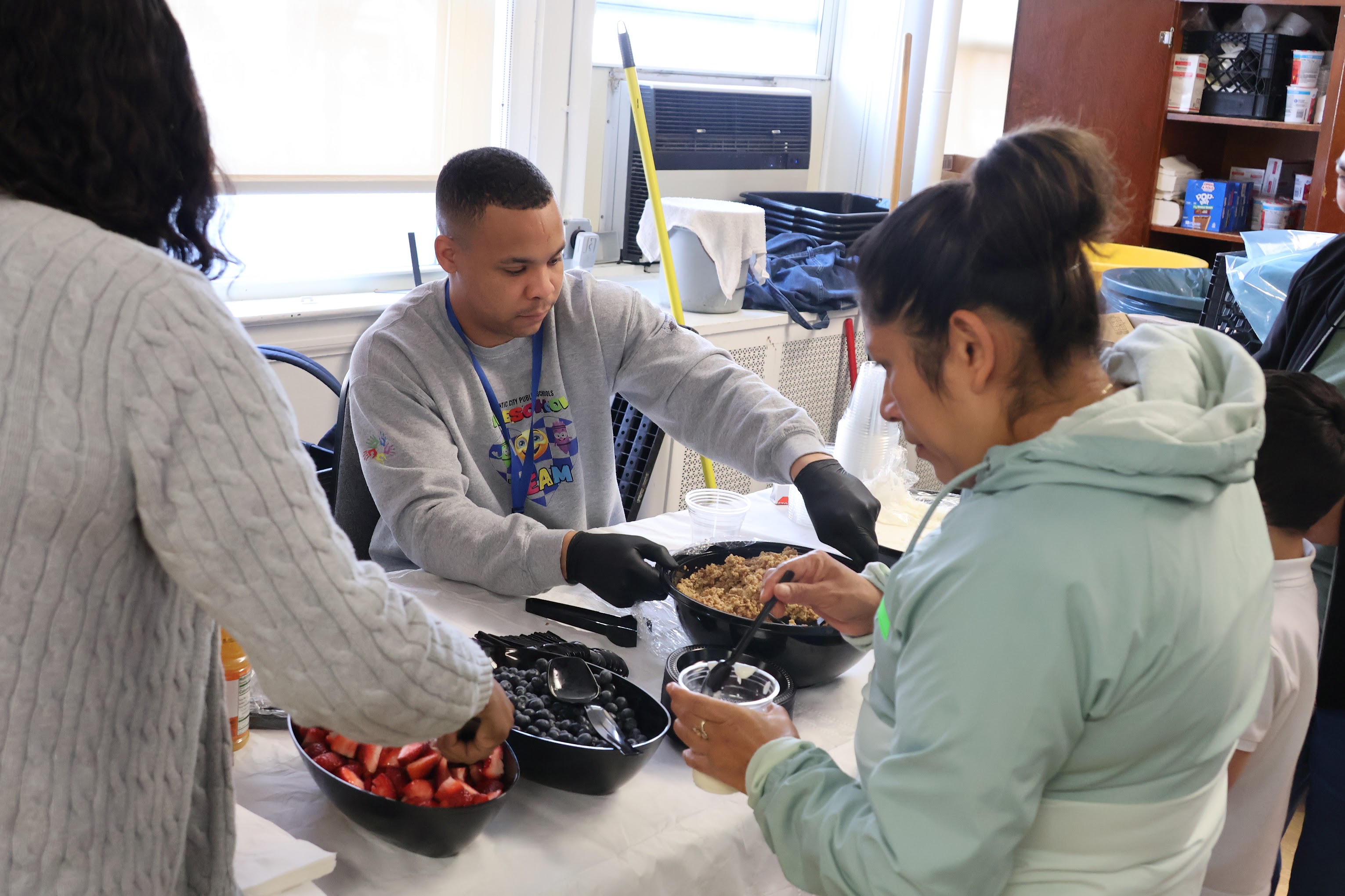 A group of people are gathered around a table, examining and discussing various food items and containers in what appears to be a kitchen or food preparation area.