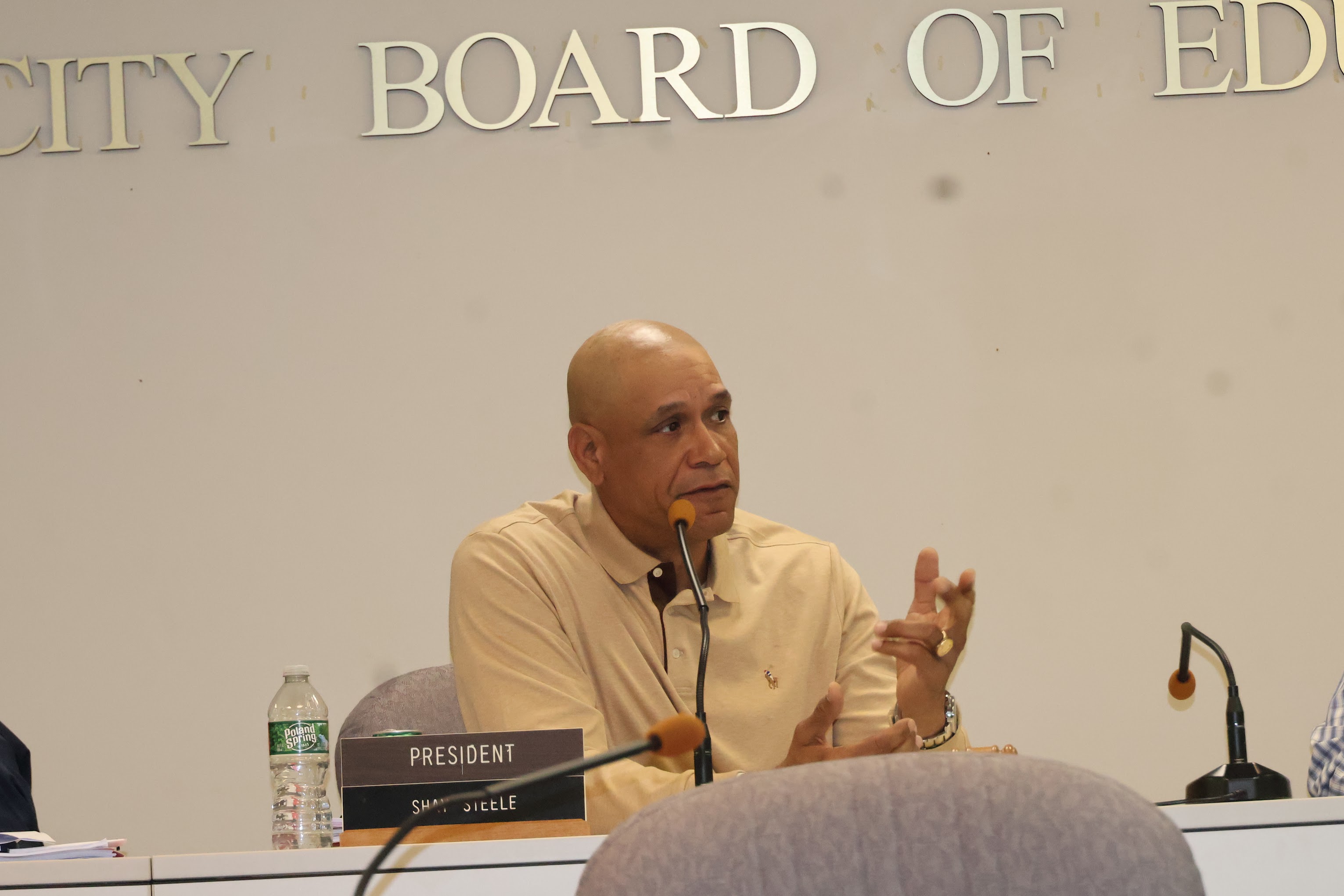 The image shows an older man, likely a member of the Board of Education, speaking at a podium in front of a sign that reads "City Board of Education".