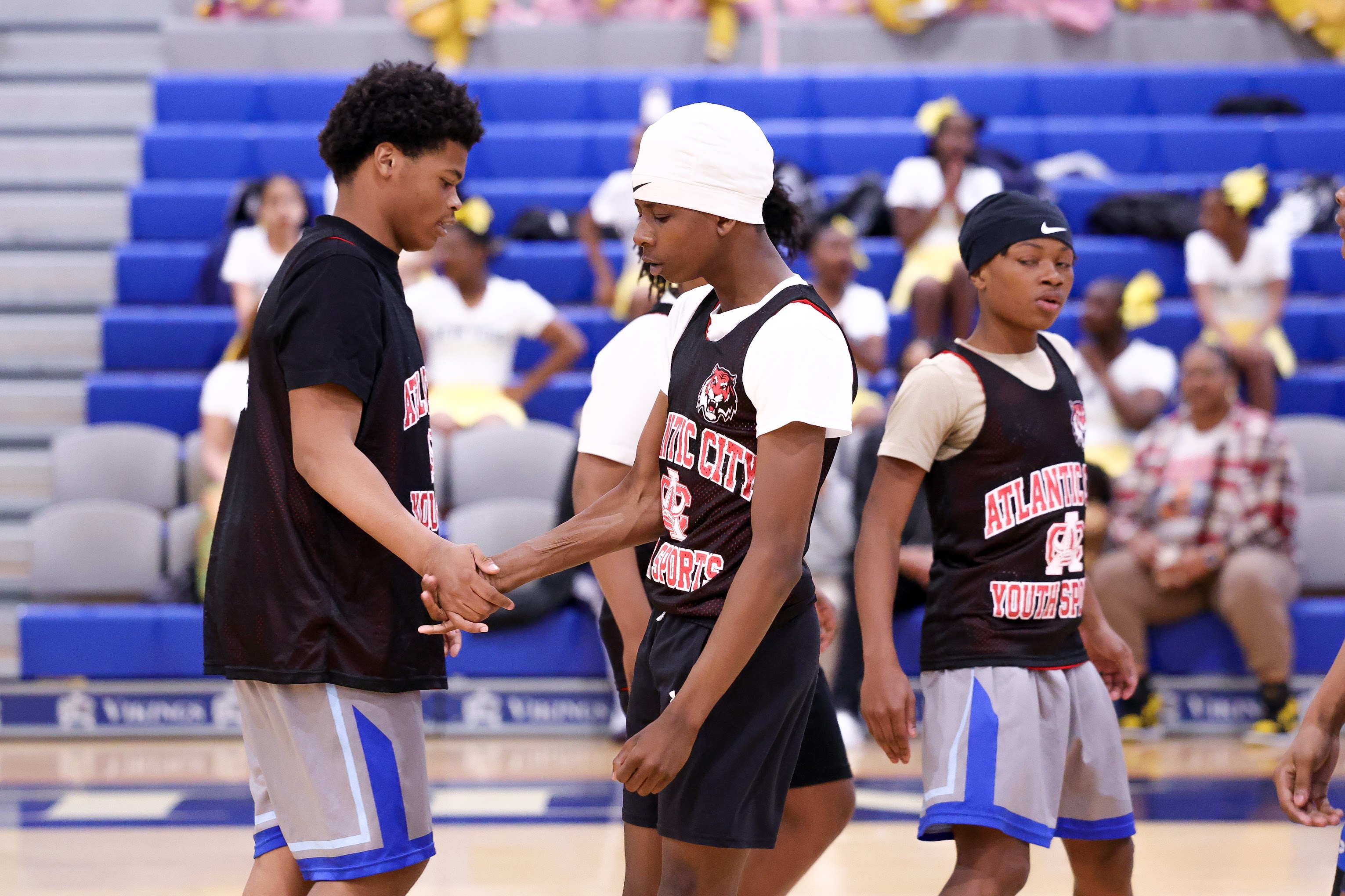 The image shows three young basketball players standing together on a basketball court, with a crowd of spectators visible in the background.
