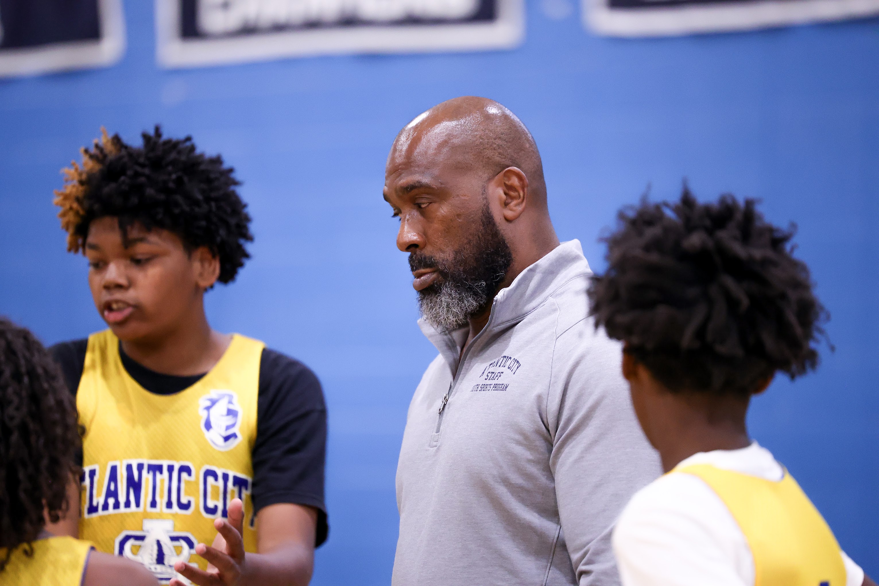 The image shows a man with a beard standing between two young people wearing yellow jerseys, with a blue background behind them.