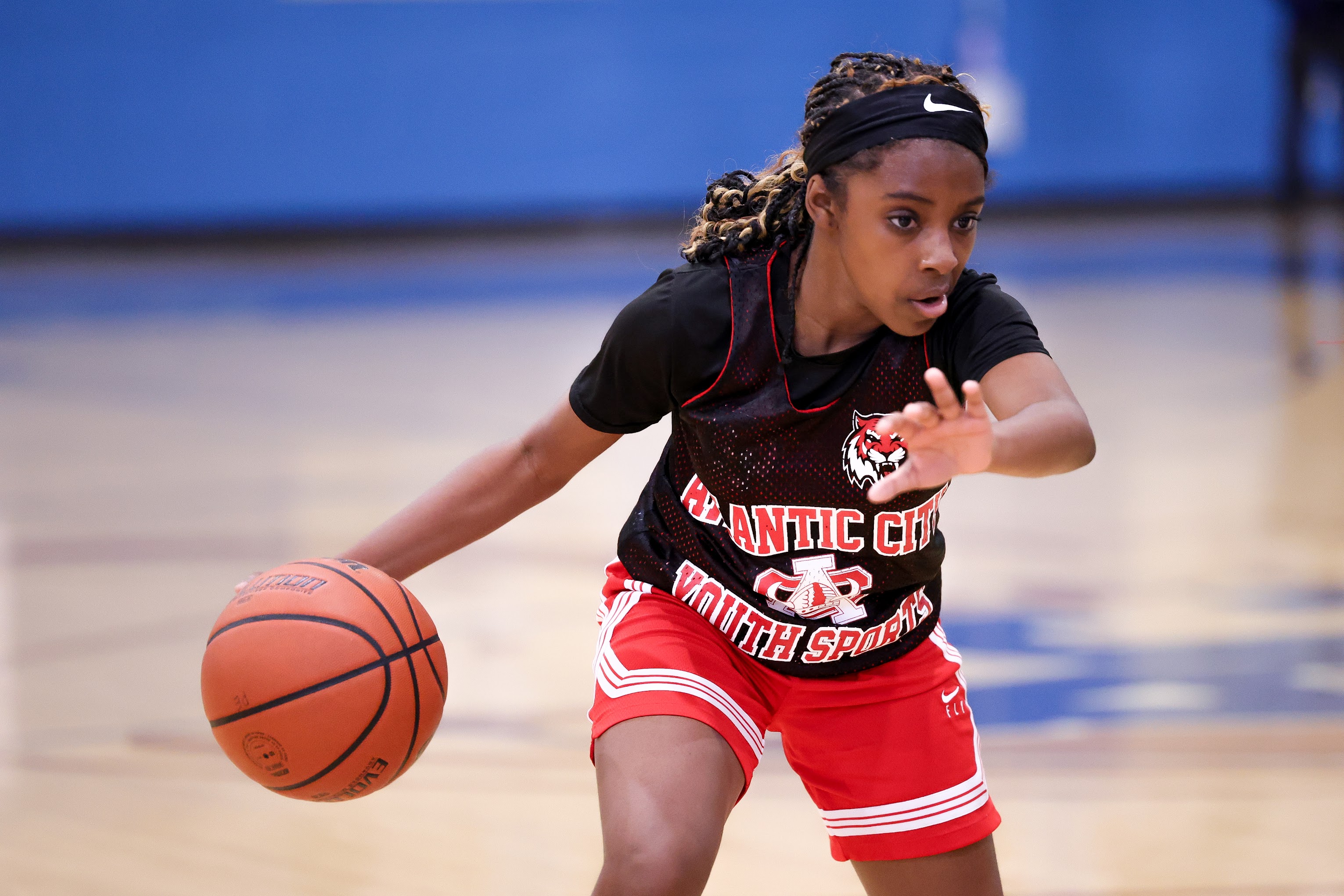 A young female basketball player in a red and black uniform dribbling a basketball on a basketball court with a blue background.