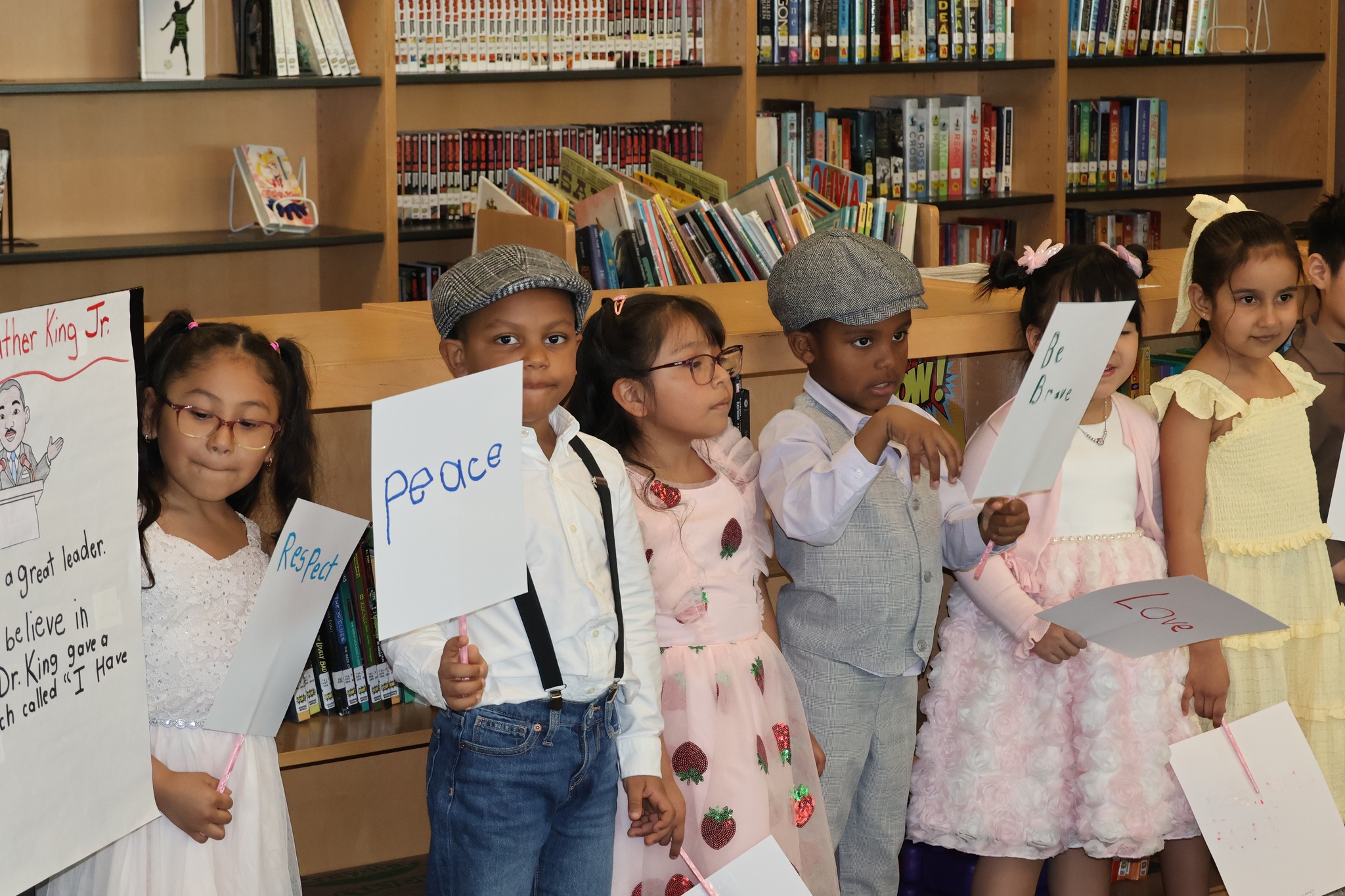 A group of young students, some holding whiteboards, stand in front of bookshelves in what appears to be a library or classroom setting.
