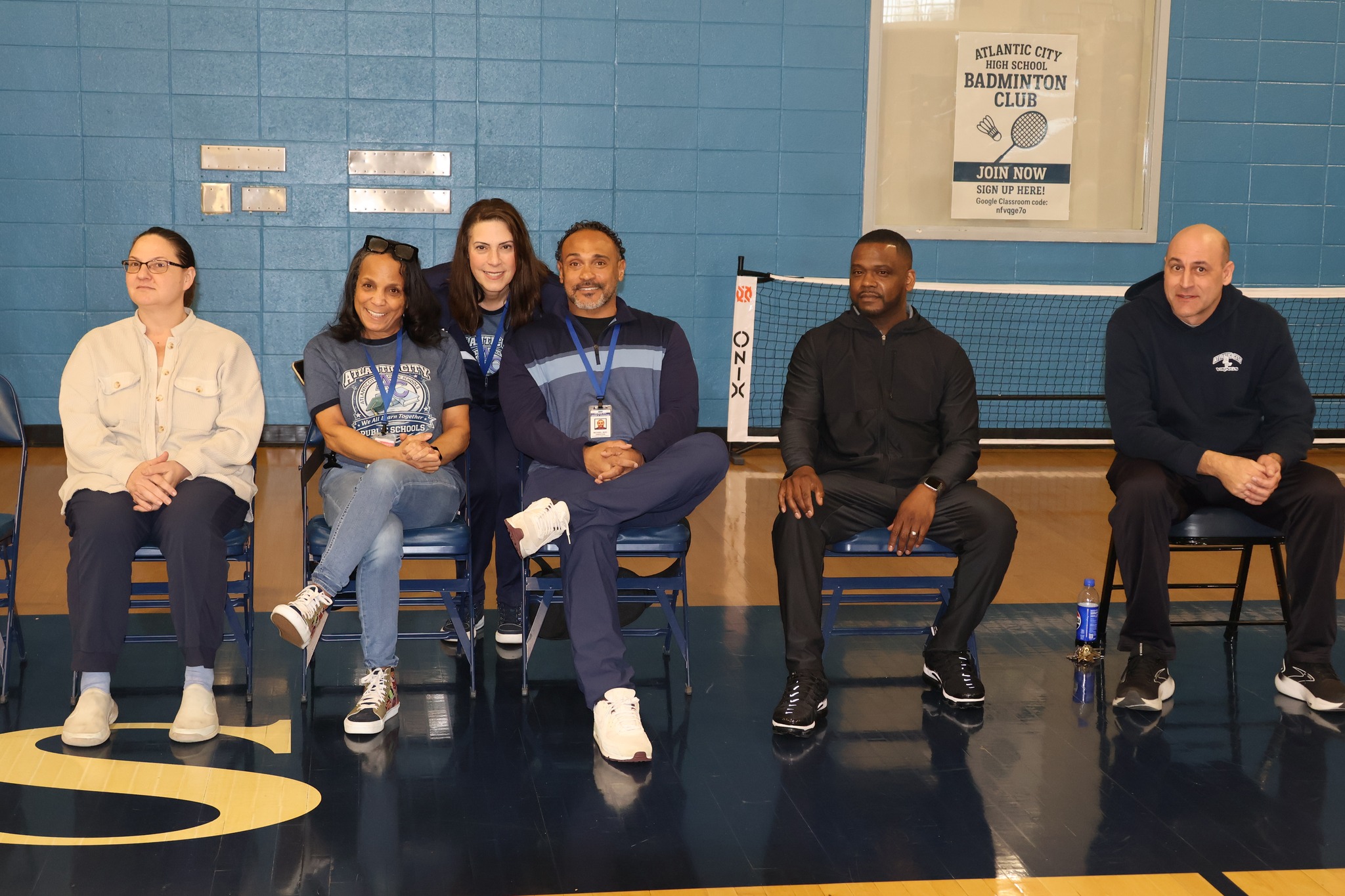 A group of people, including both men and women, are seated in a gymnasium-like setting with a badminton court visible in the background.