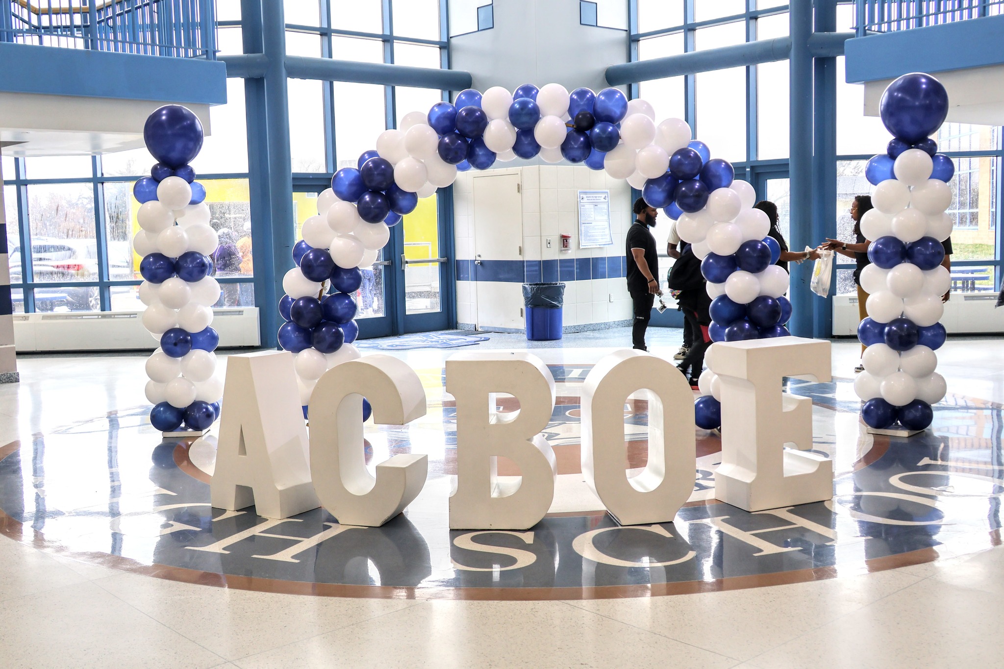 The image depicts a large, decorative display featuring the word "ACROS" made of white and blue balloons, set against a backdrop of glass windows and a bright, open interior space.