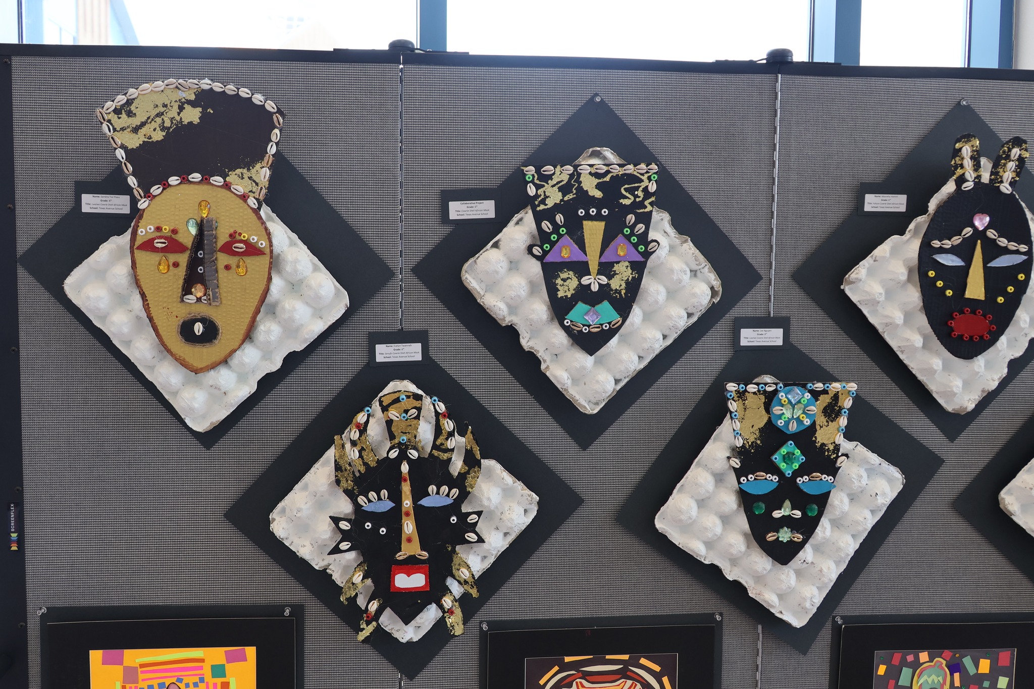 Colorful, decorative masks with intricate designs are displayed on a gray wall with diamond-shaped backings, while framed artwork is visible in the foreground.
