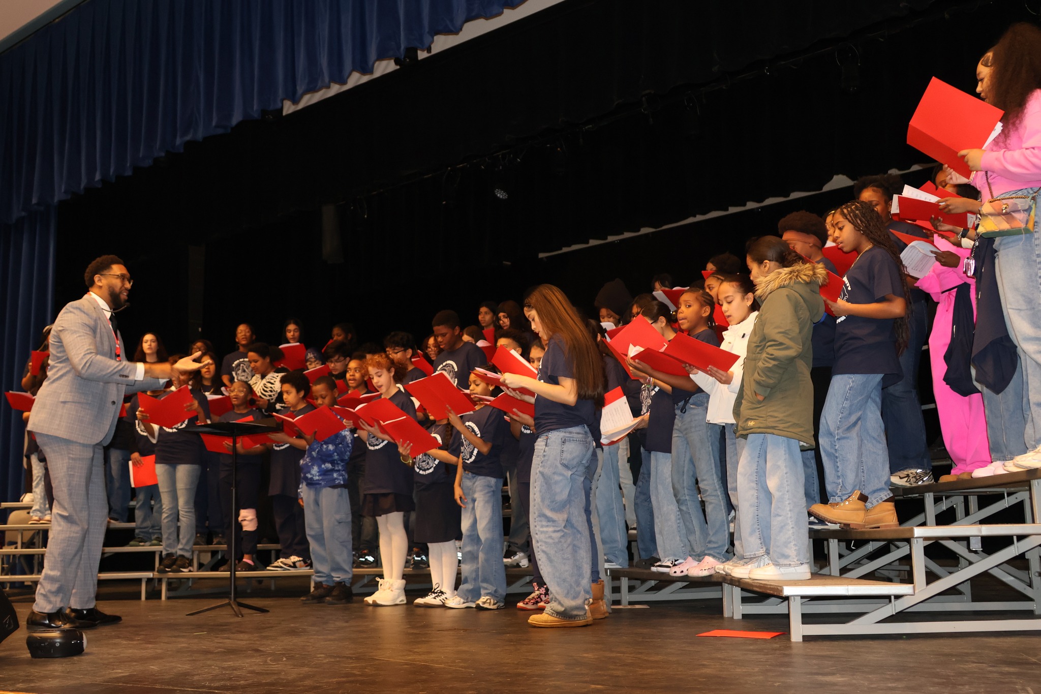 A choir of students in blue shirts and jeans, holding red songbooks, stands on risers on a stage, directed by a man in a suit.