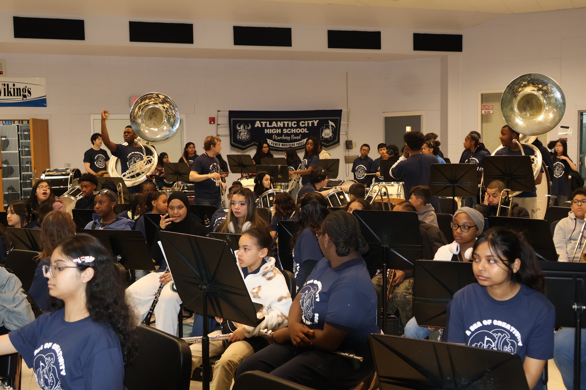 A high school band, with students in the foreground playing various instruments, performs in a room with a banner reading "Atlantic City High School" in the background.