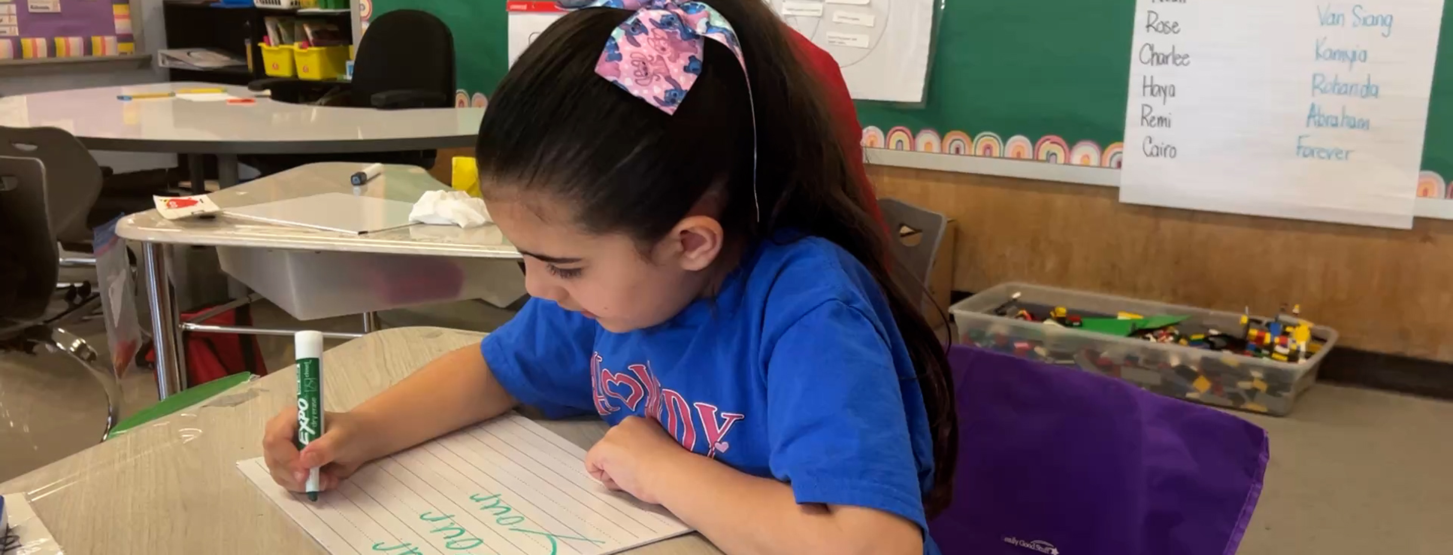 A young girl with a floral bow in her ponytail writes on a paper at her desk, with a bin of colorful building blocks and a whiteboard visible in the classroom background.