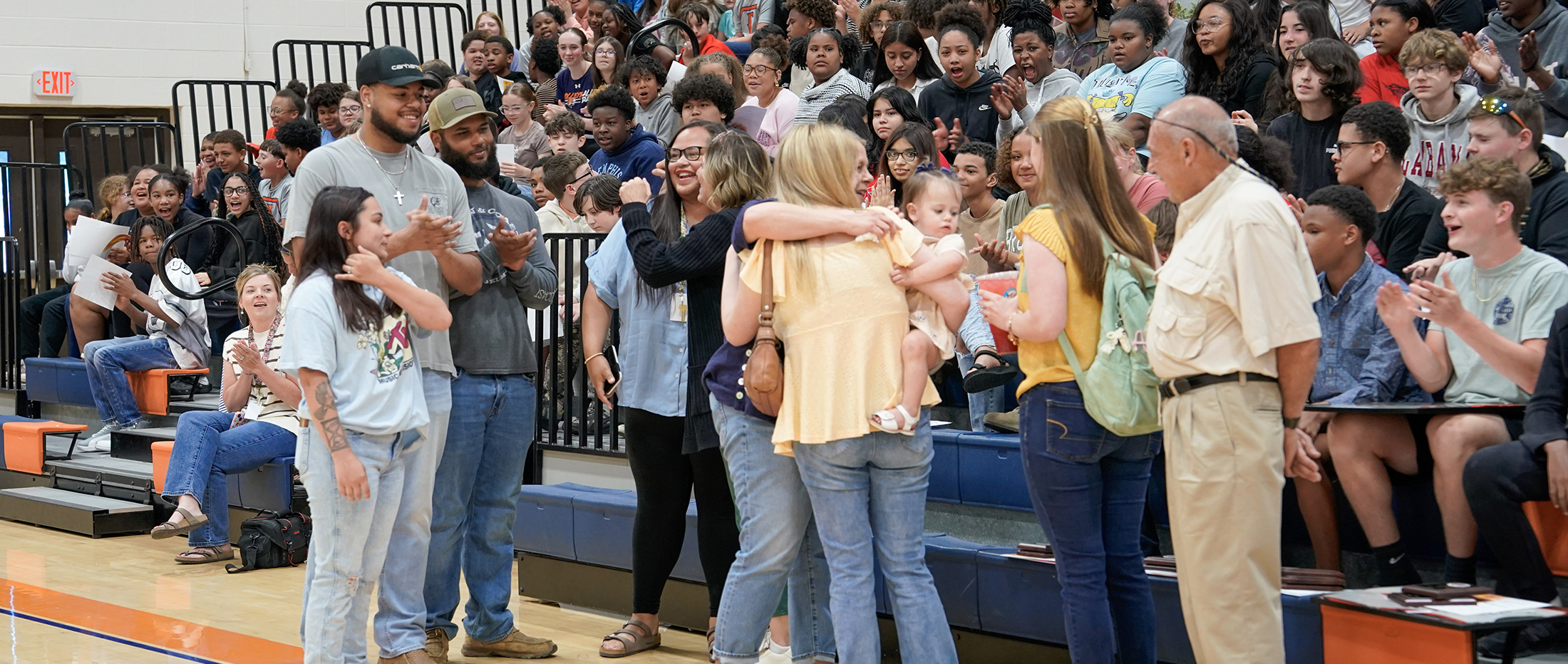 In the foreground, a group of people are gathered, some hugging and holding a baby, while in the background, a large crowd of spectators watches from bleachers.