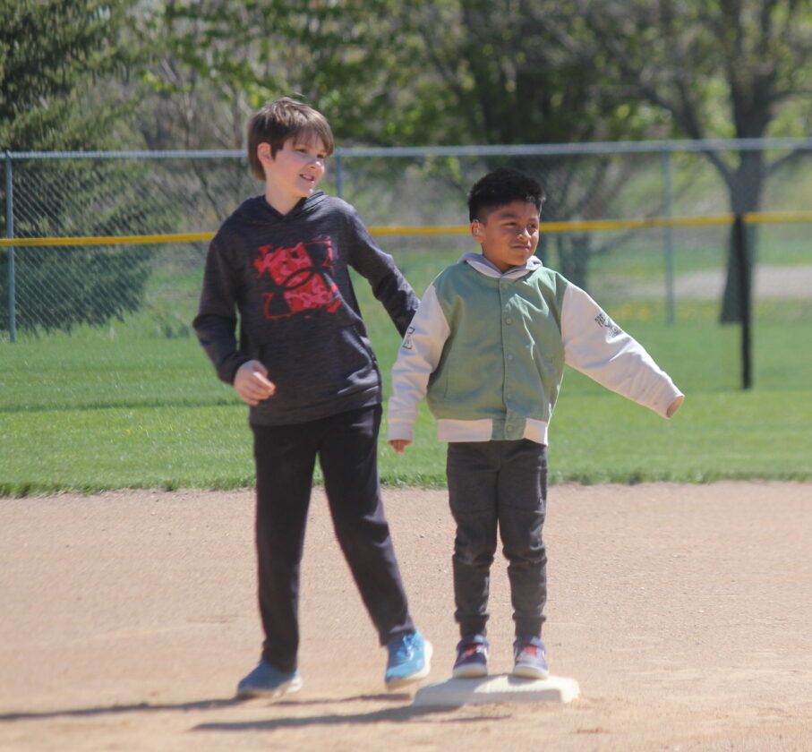 Two young boys stand on a baseball field with a fence and trees in the background.