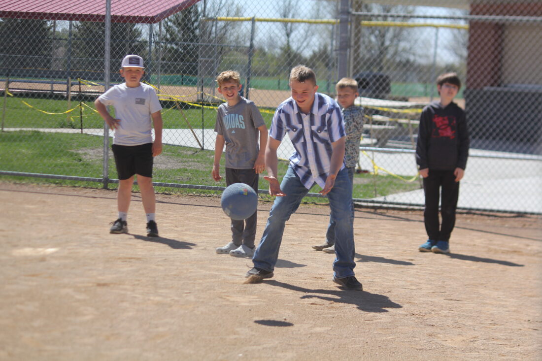 Five boys play kickball on a dirt field with a fence and trees in the background.