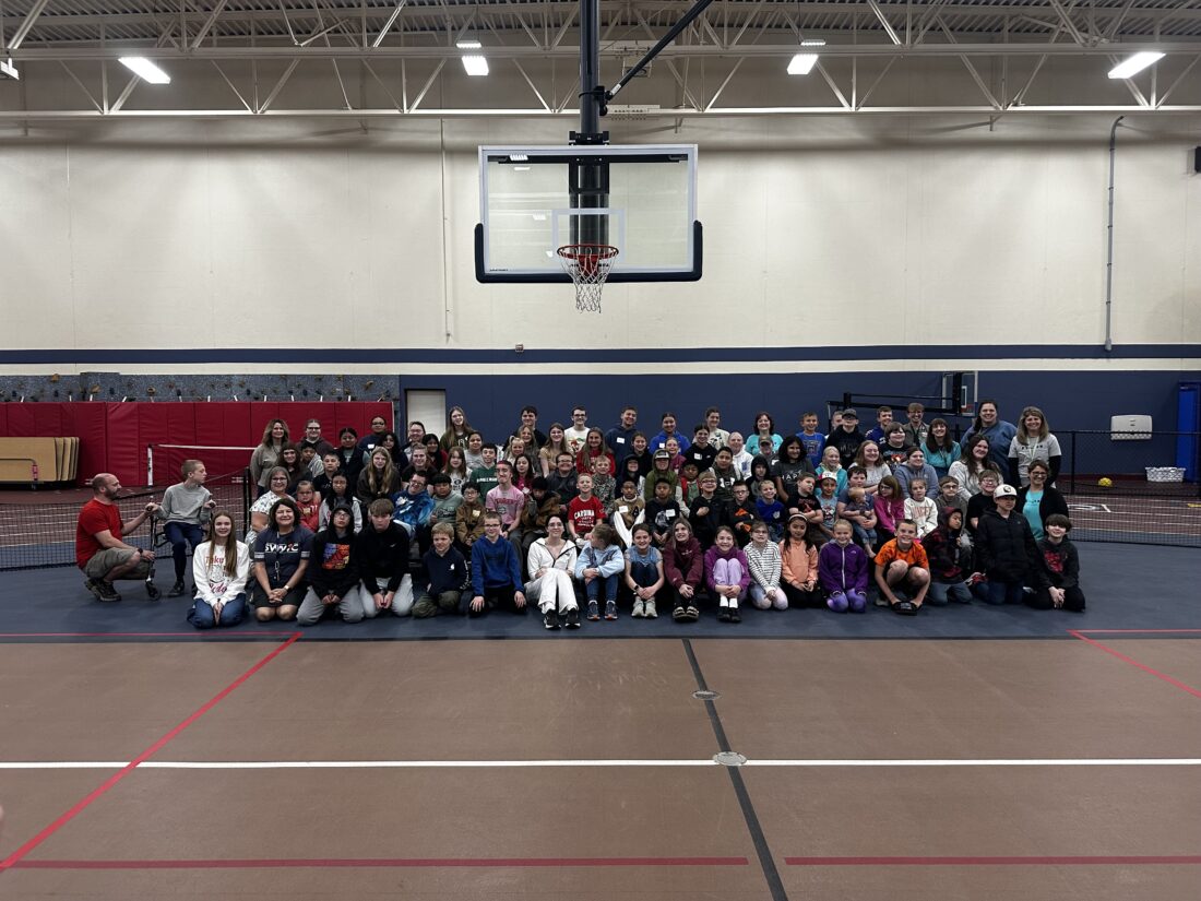 A large group of people, including children and adults, are posing for a photo on a basketball court with a basketball hoop visible in the background.