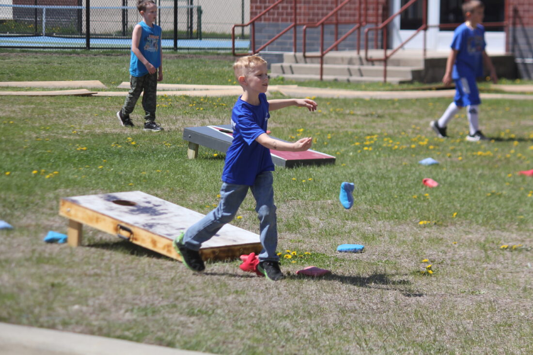 A young boy in a blue shirt throws a beanbag at a cornhole board in the foreground, while other children play in the grassy background near a brick building.