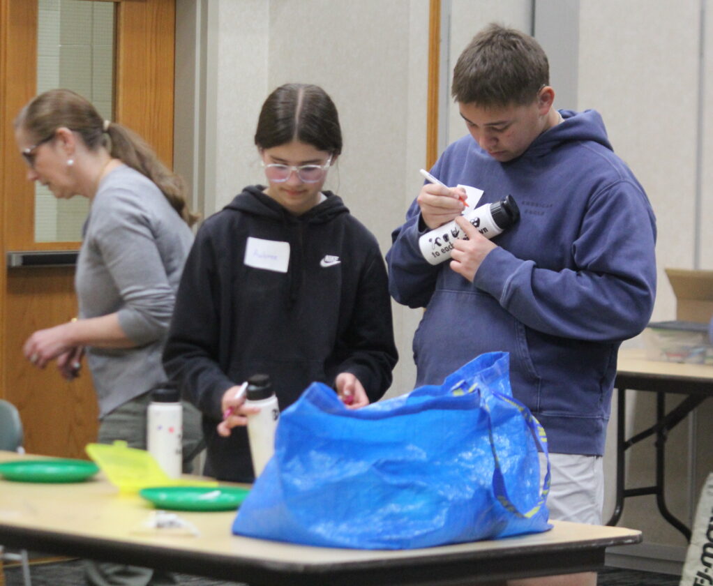 In the foreground, two young people are personalizing water bottles at a table with a blue bag, while in the background, an adult woman stands near a doorway.
