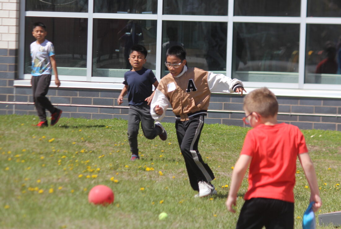 Four boys play on a grassy field in front of a brick building with large windows.