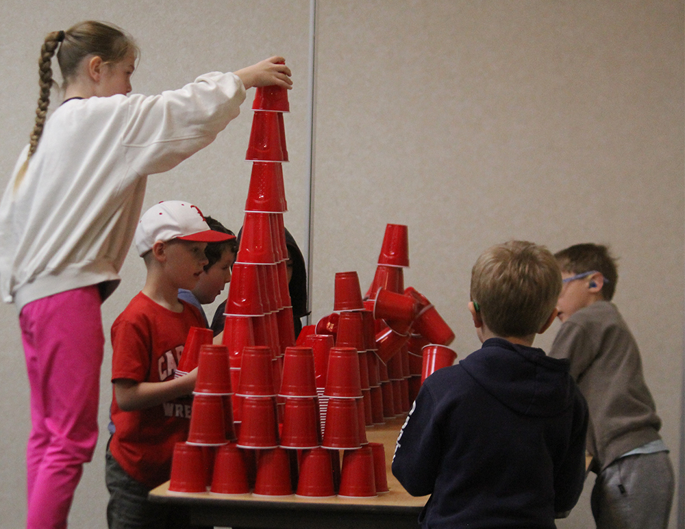 Children in the foreground build a tall tower of red cups, while a plain wall forms the background.