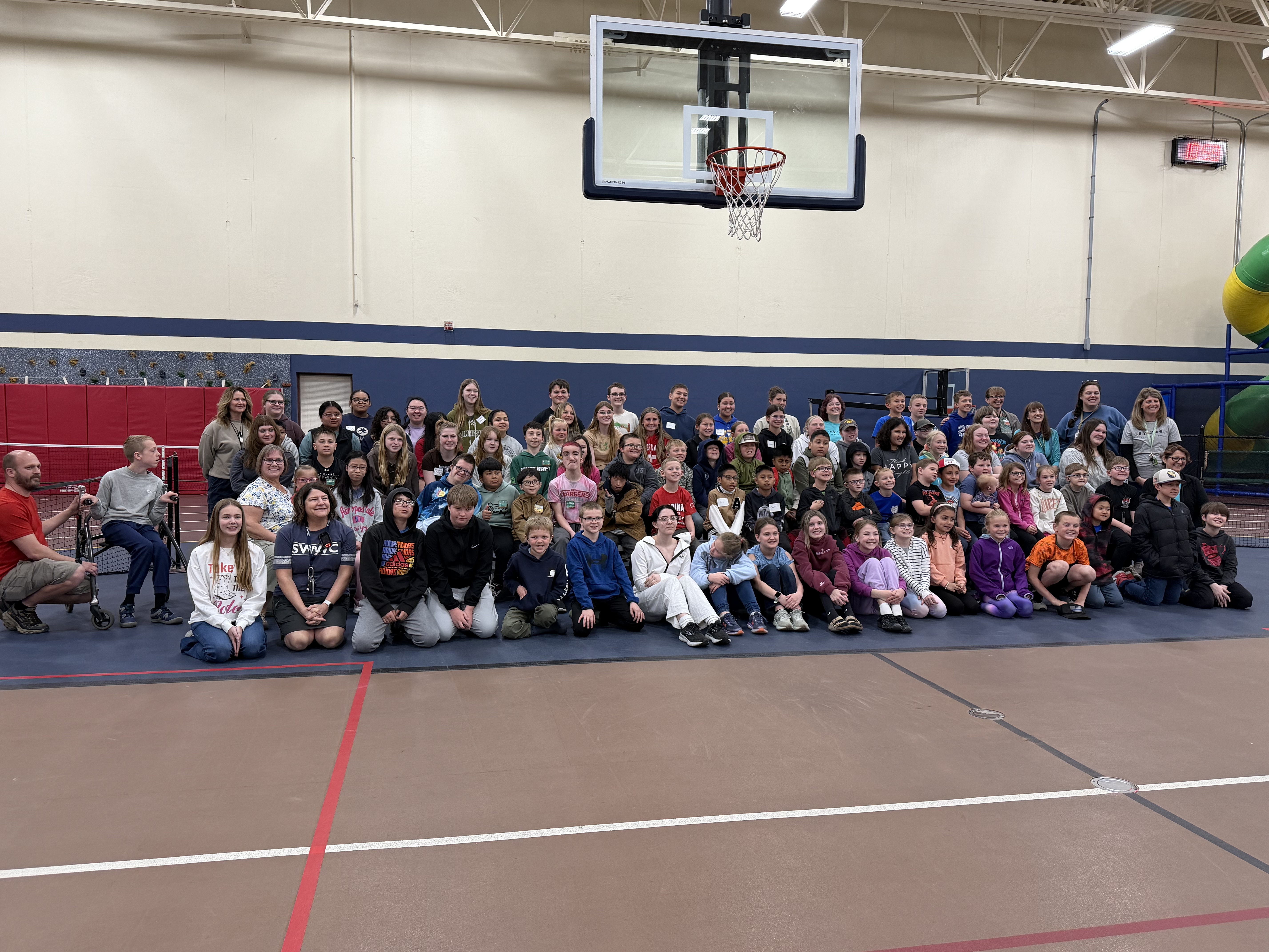 A large group of people of all ages are posing for a photo in a gymnasium with a basketball hoop in the background and a red line on the floor in the foreground.