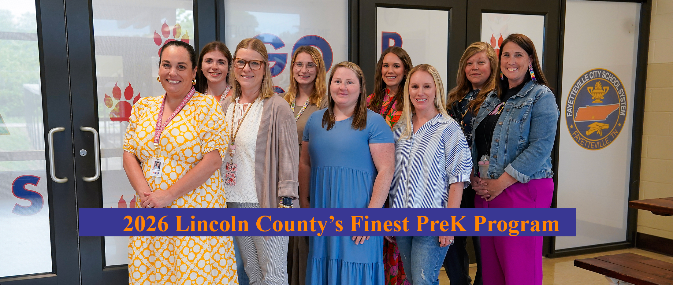 A group of nine women stand in front of a school entrance with a banner that reads "2026 Lincoln County's Finest PreK Program."