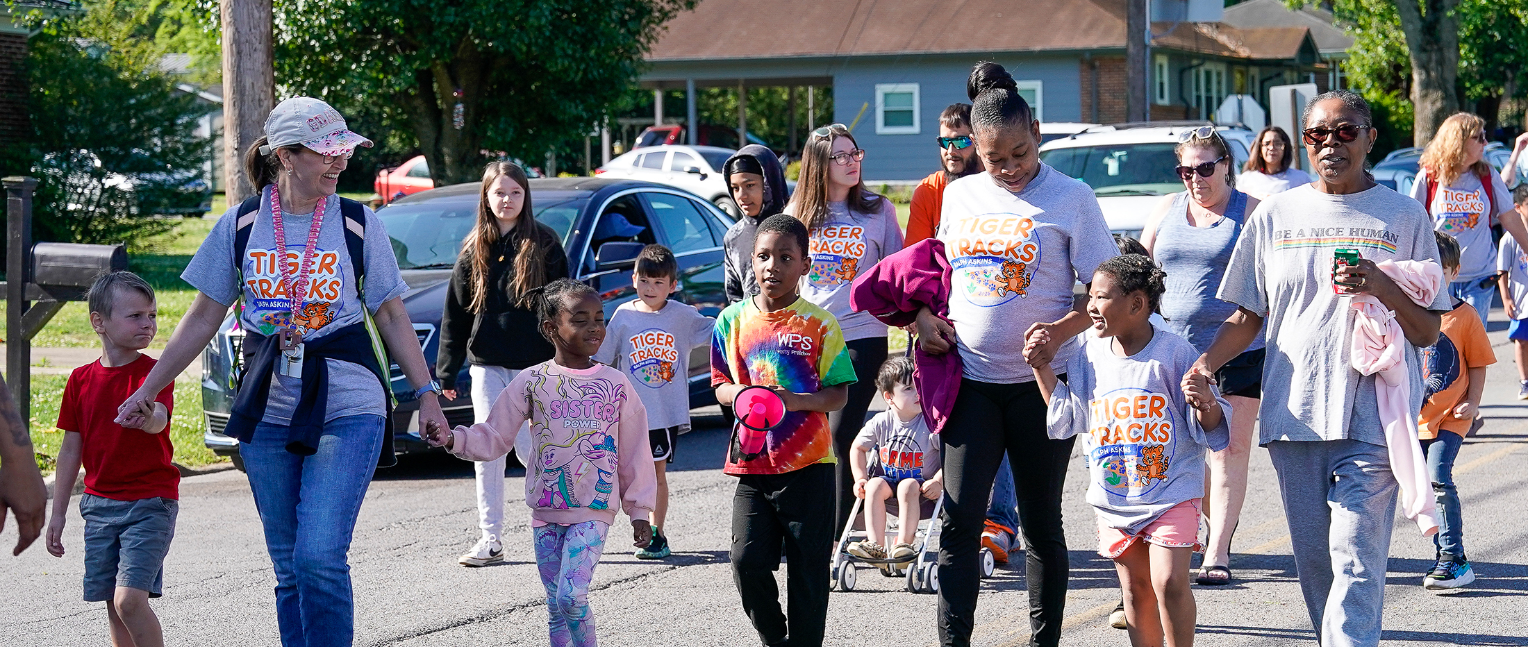A group of people, including children and adults, walk down a street in a parade, with houses and cars visible in the background.