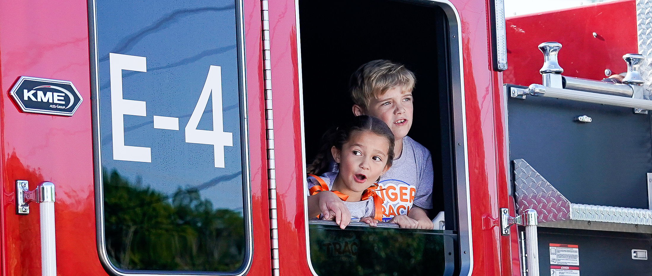 Two excited children peer out of the window of a red fire truck with "E-4" visible on its side, against a blurred background of trees and sky.
