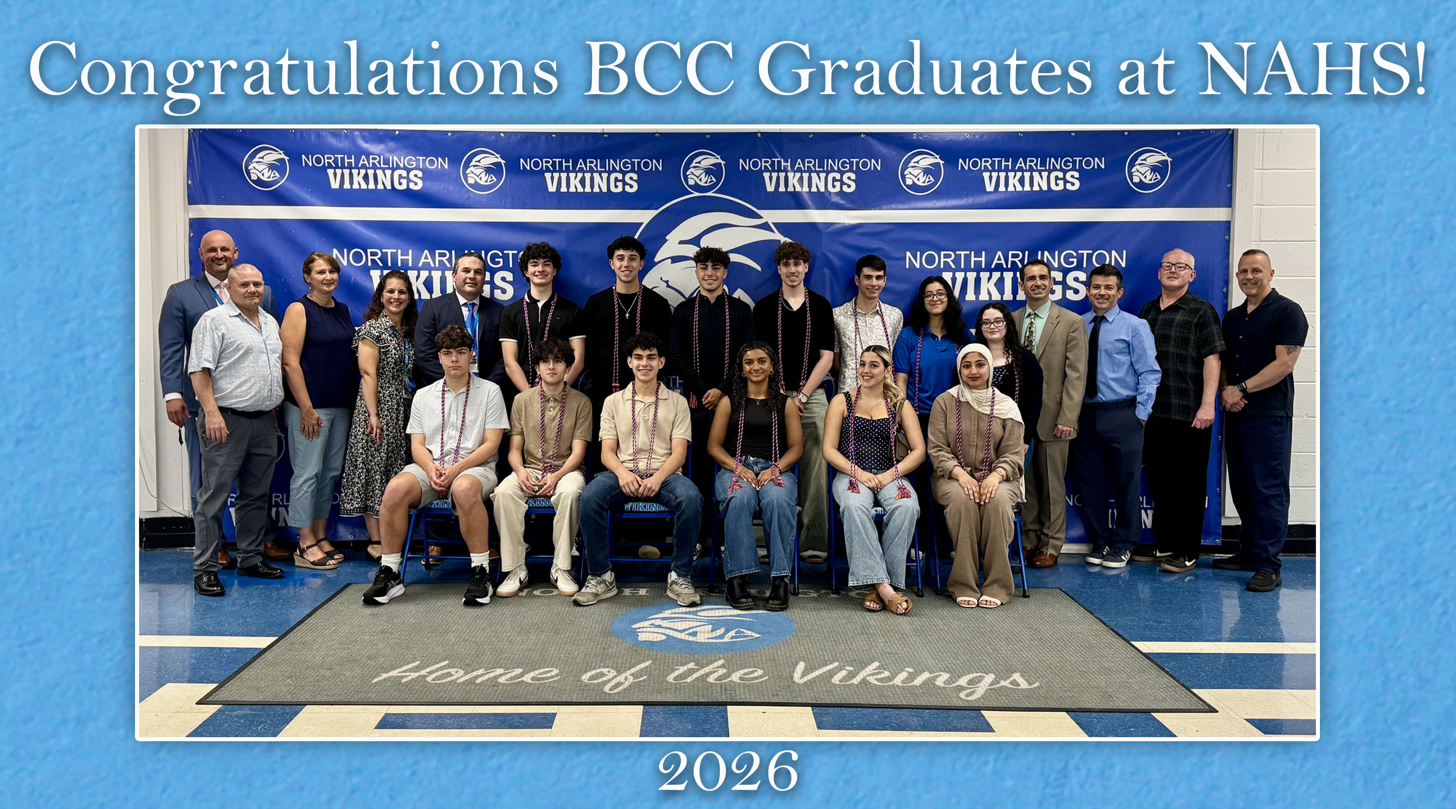 A group of graduates and staff pose for a photo in front of a banner that reads "Congratulations BCC Graduates at NAHS!" with "Home of the Vikings" and the year "2026" visible on the floor.