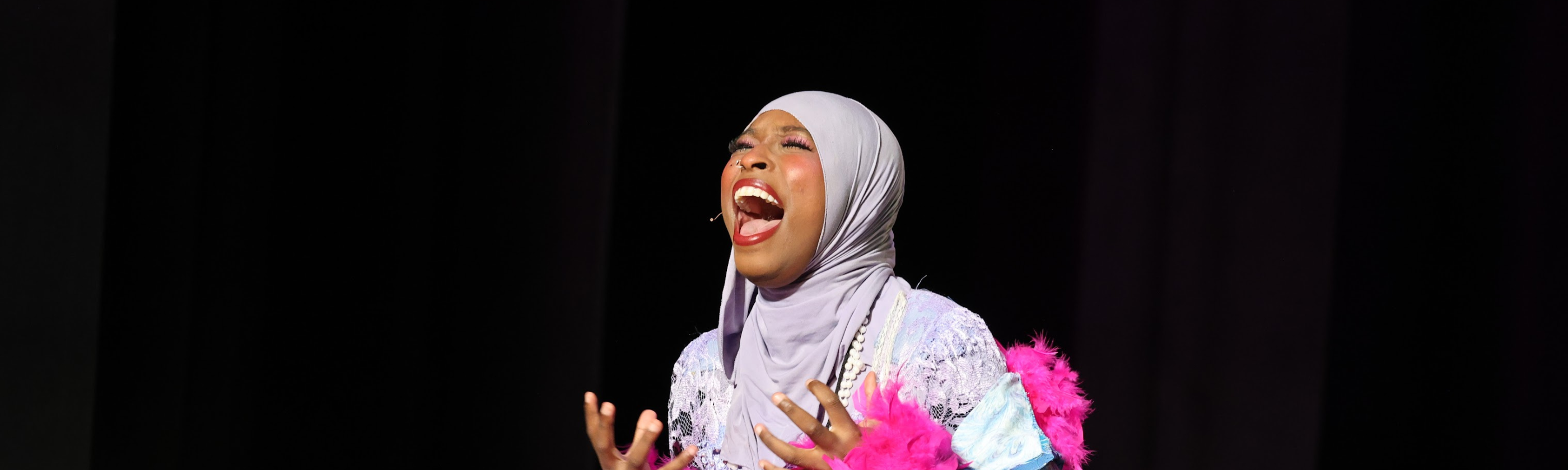 A woman in a light purple hijab and a sparkly, feathered costume belts out a song on a dark stage.