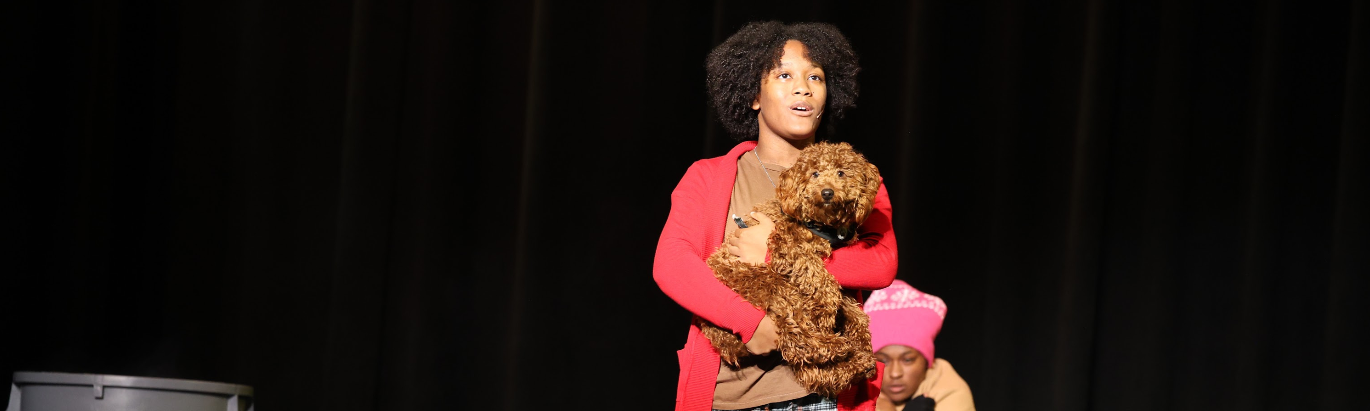 A young person in a red cardigan holds a fluffy brown stuffed dog on a dark stage, with another person in a pink hat visible in the lower right corner.