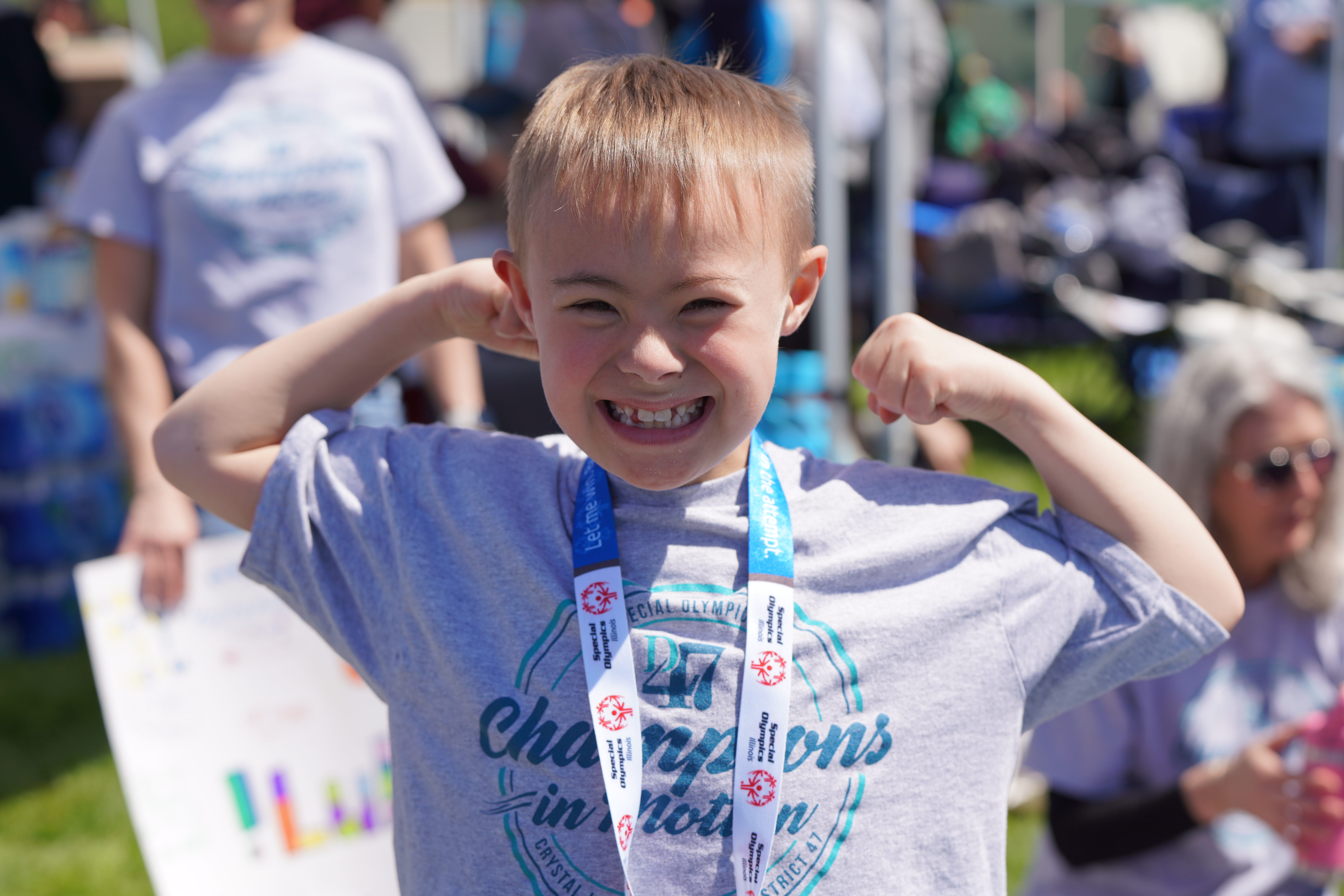 A young boy proudly flexes his muscles in the foreground, wearing a medal, while blurred figures and tents form the background of an outdoor event.
