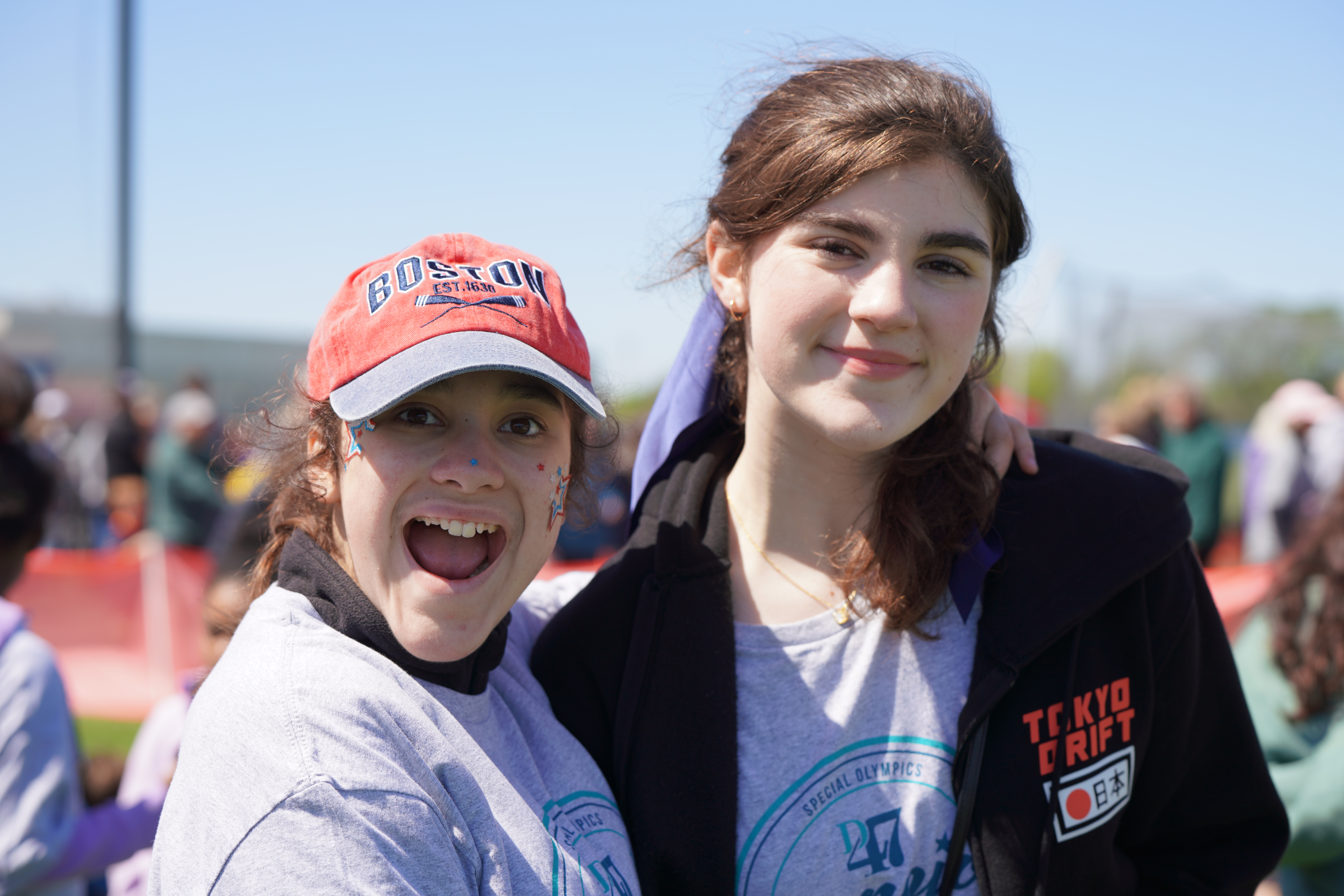 Two young women smile at the camera in the foreground, with a blurred crowd and a bright sky in the background.