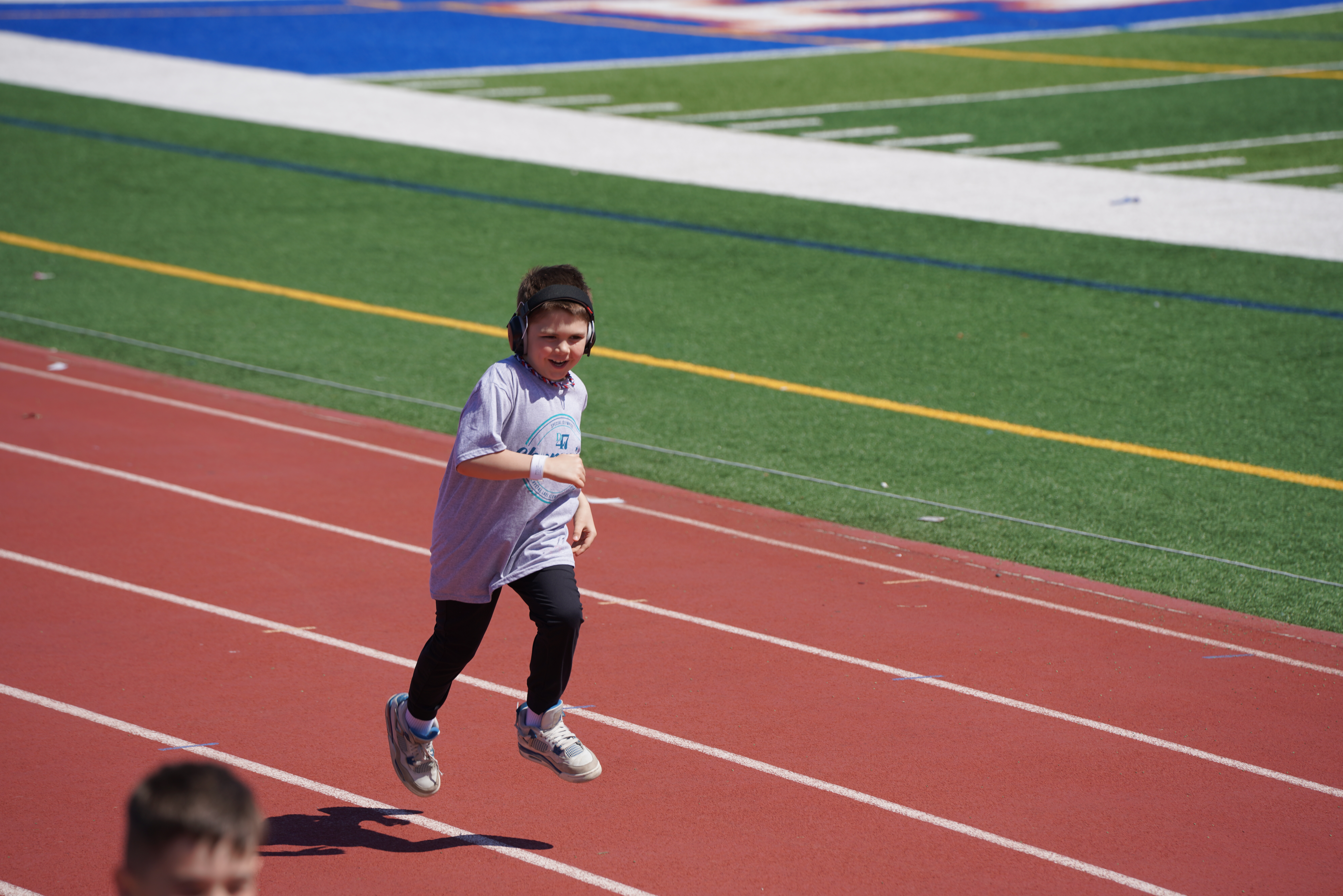 A young boy in headphones runs on a red track in the foreground, with a blurred person and a green and blue field in the background.