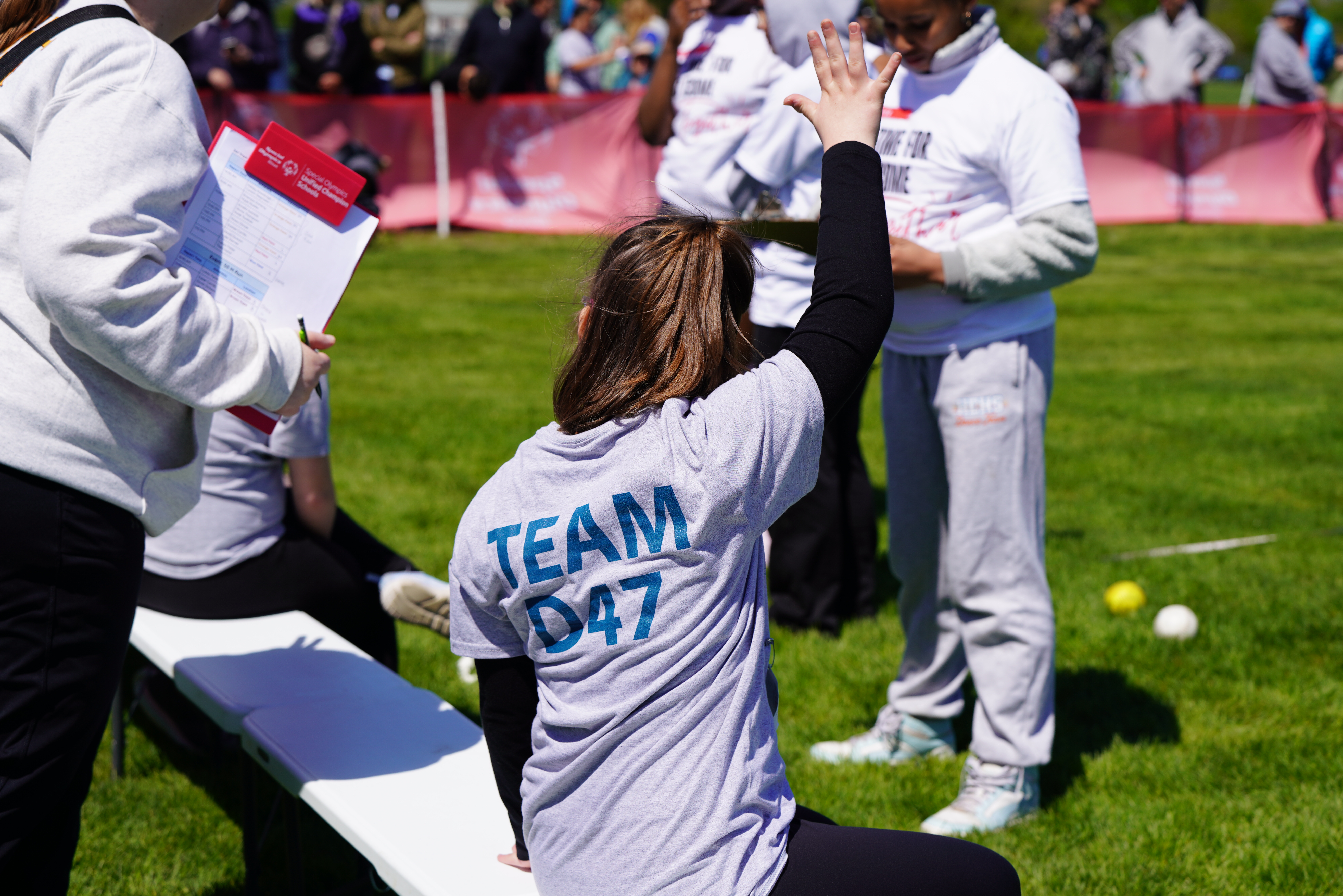 In the foreground, a girl in a "TEAM D47" shirt gives a high-five, while in the background, spectators watch an outdoor event.