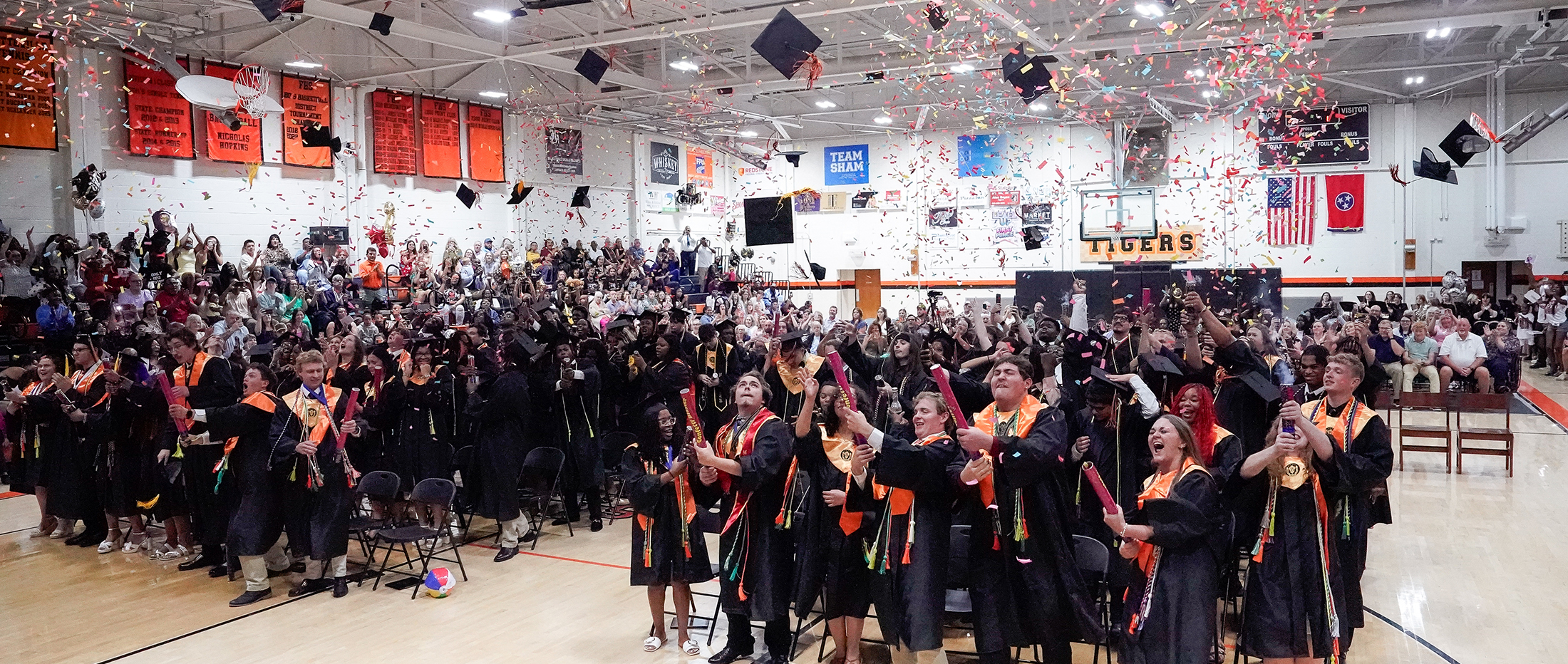 Graduates in black gowns with orange sashes cheer and throw confetti in a gymnasium filled with spectators.